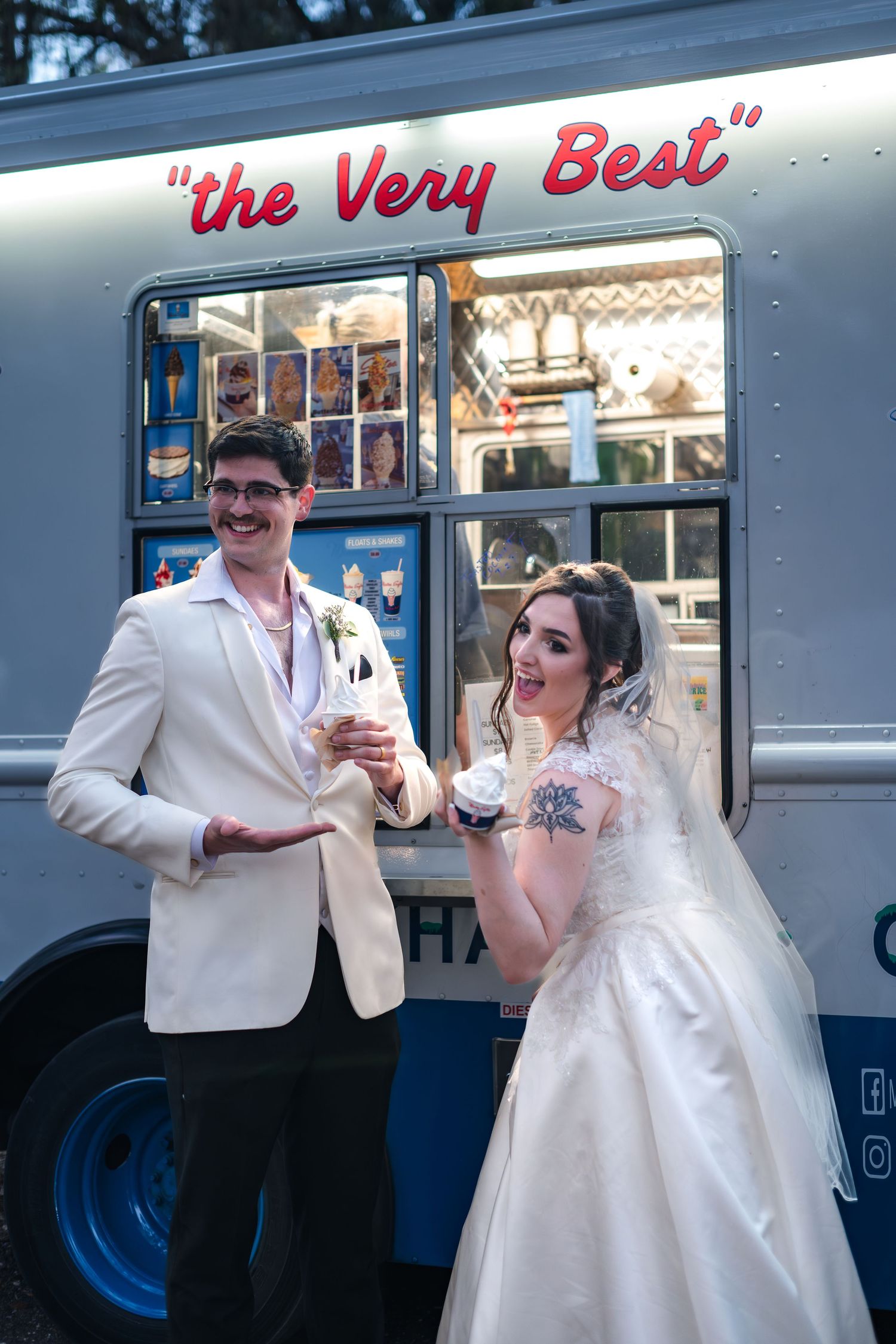 A couple in wedding attire poses while enjoying drinks from a vintage food truck with 'the Very Best' signage.