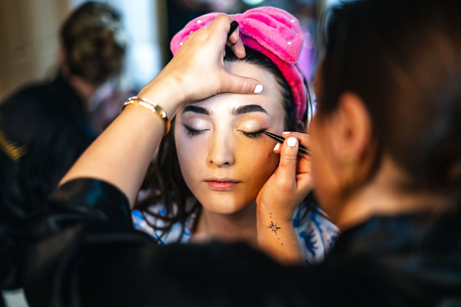 Makeup artist applies eye makeup while person wears a bright pink cap during a beauty preparation session.