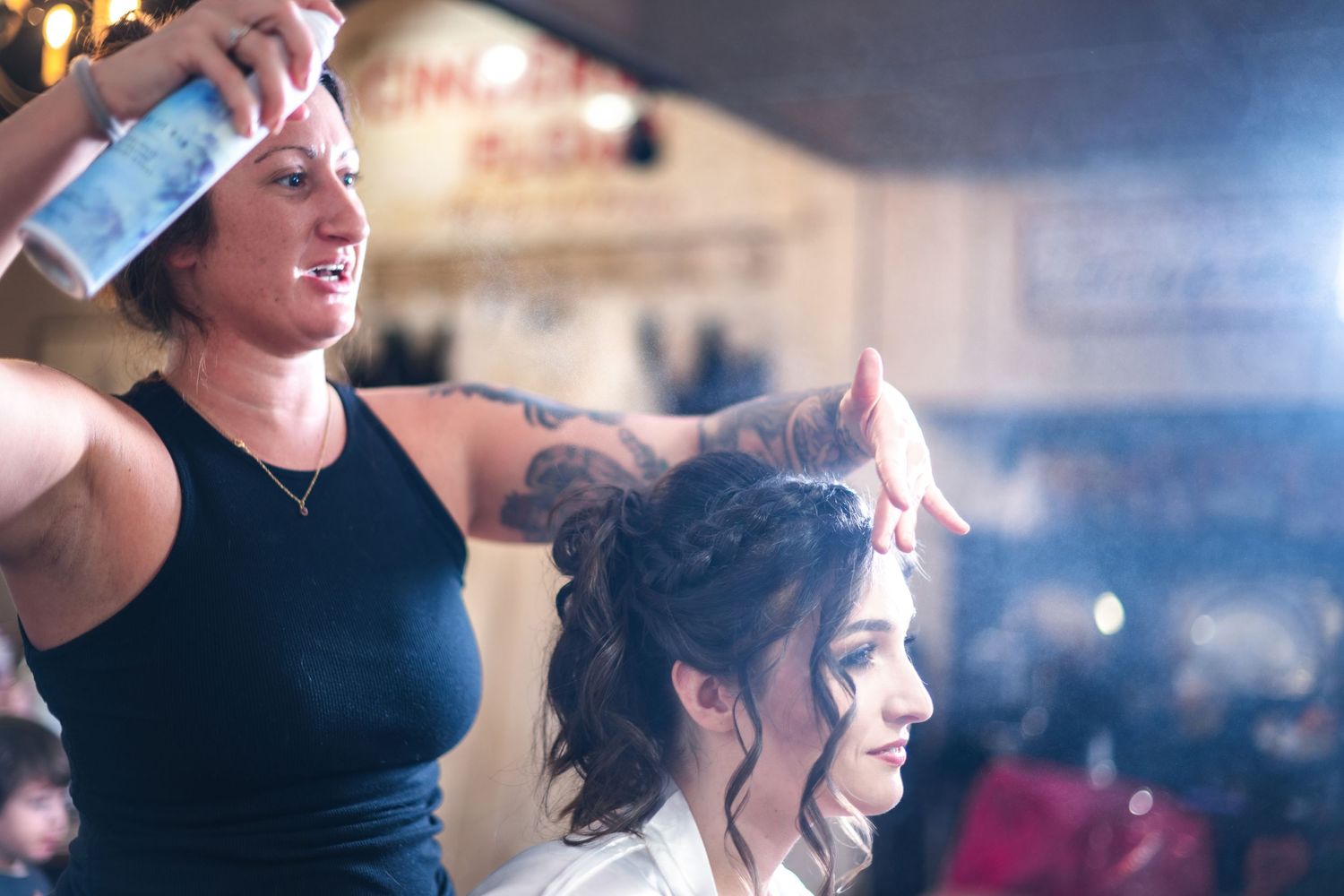 A hairstylist applies hairspray while styling hair at a salon with blurred lighting in the background.