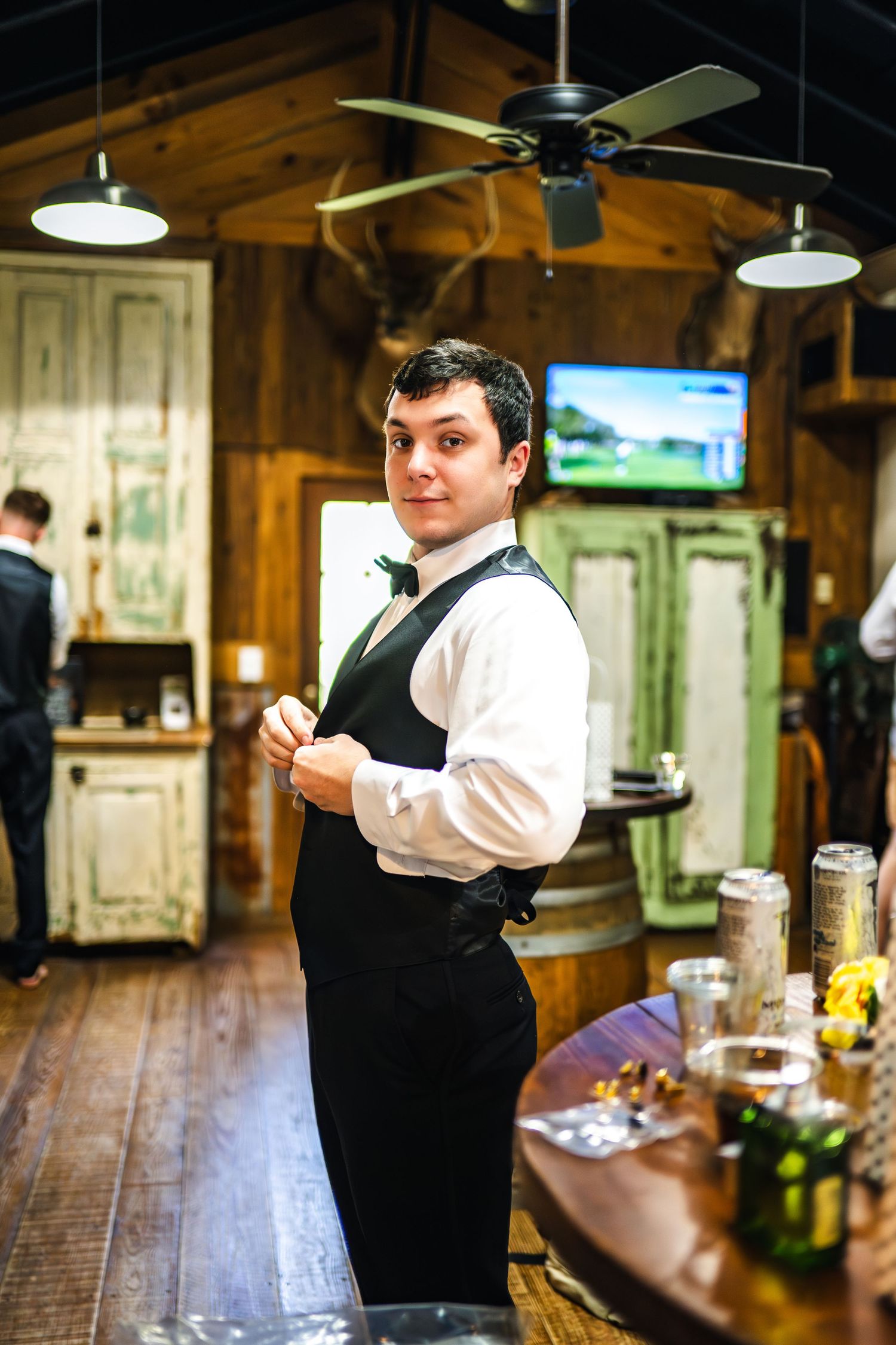 A server in formal attire stands at a rustic wooden bar beneath ceiling fans and pendant lights in a warm-lit establishment.