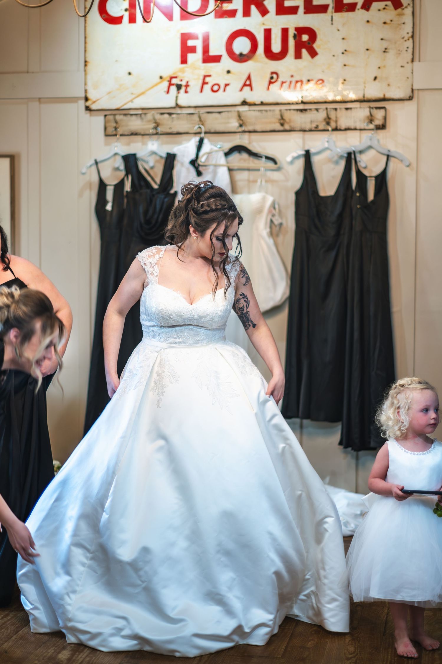 Wedding dress fitting in rustic room with vintage Cinderella Flour sign and black bridesmaid dresses hanging on wall.