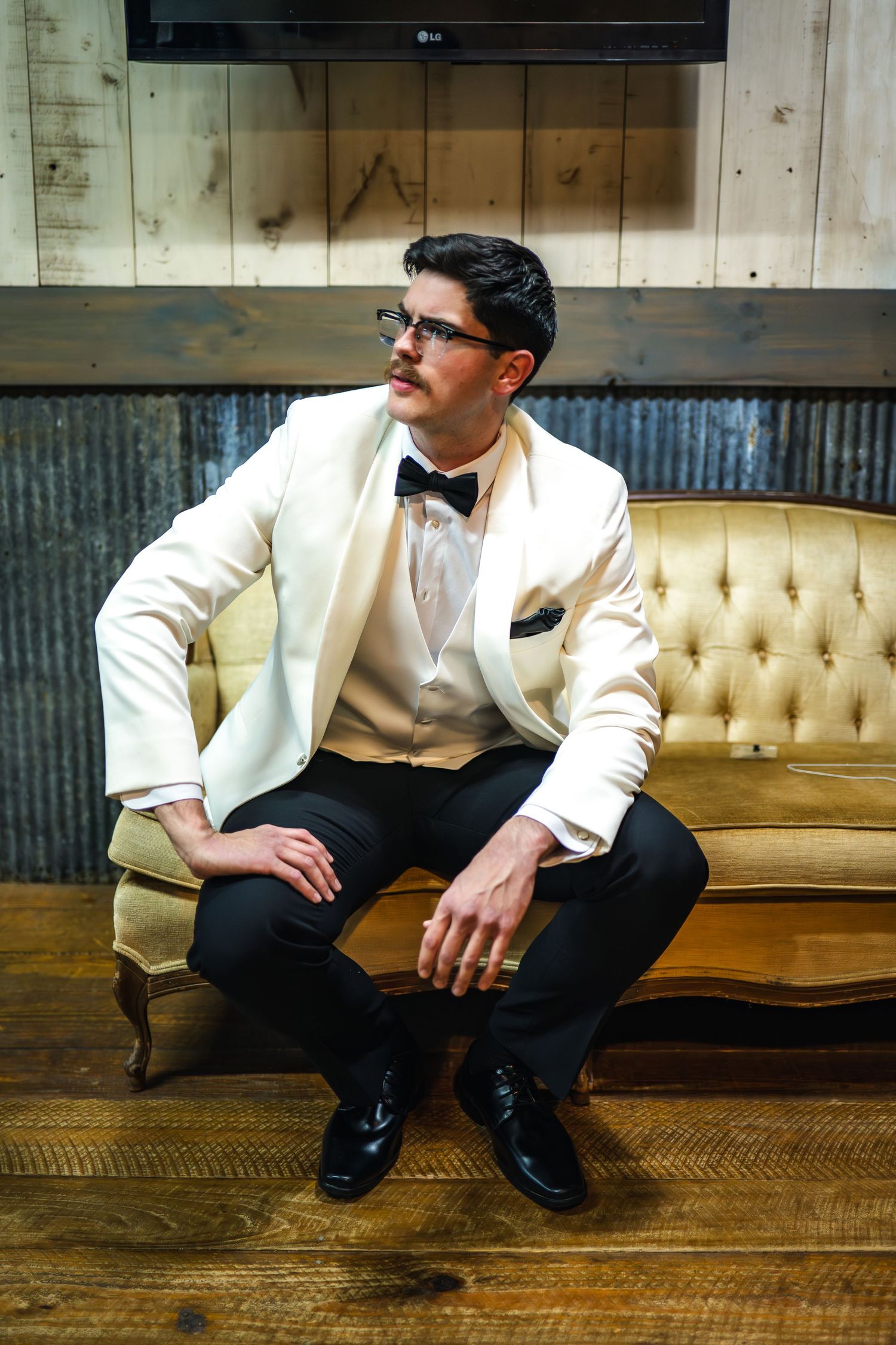 Dapper gentleman in cream tuxedo jacket and bow tie sits thoughtfully on vintage tufted leather couch against rustic wall.