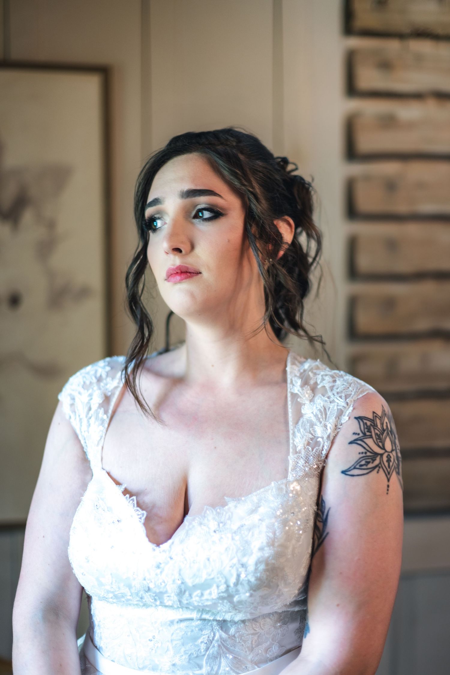 Beautiful bride in white lace wedding dress with shoulder tattoo poses against brick wall interior.