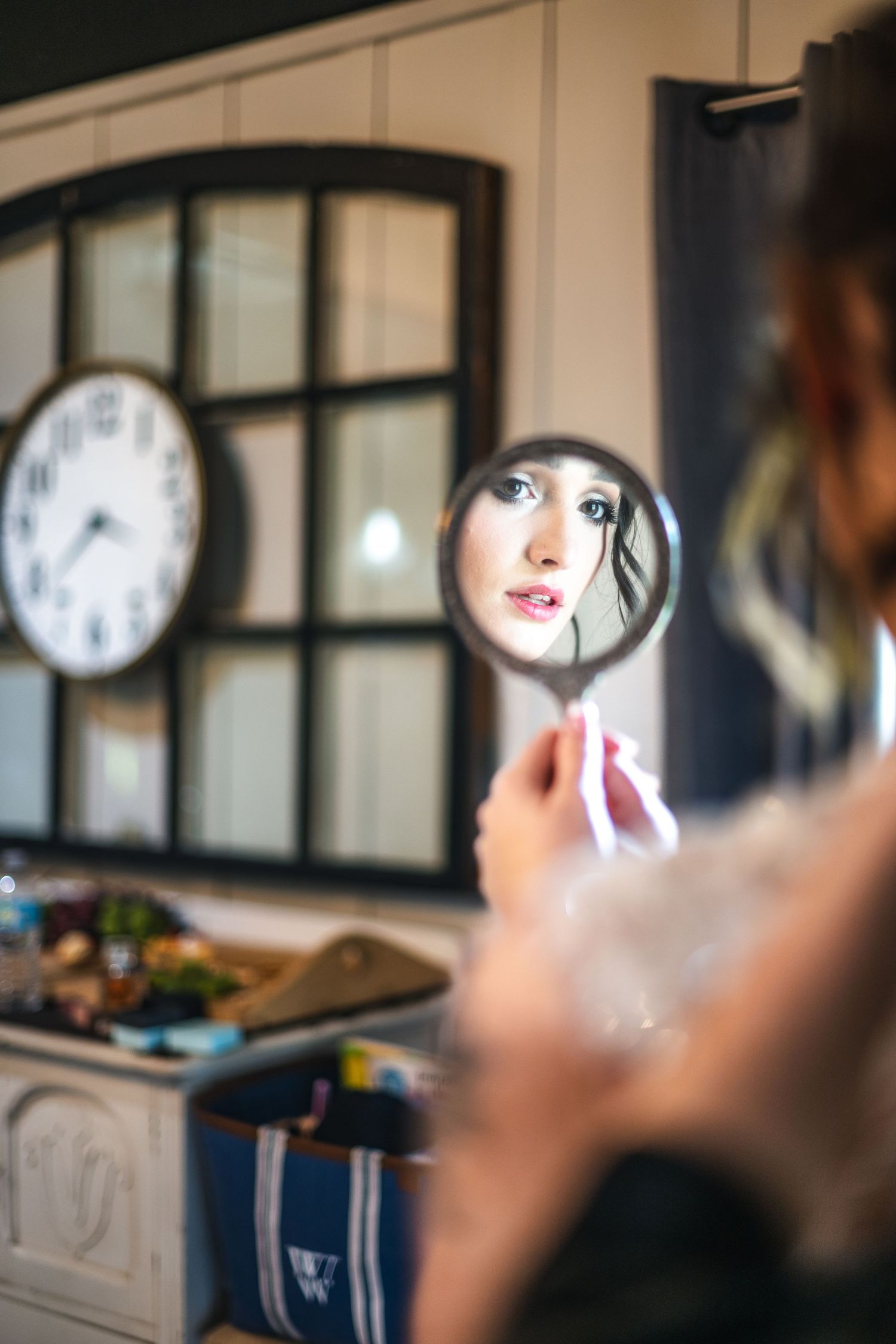 A reflection of someone's face shown in a small handheld mirror with a clock visible on the wall in the background.