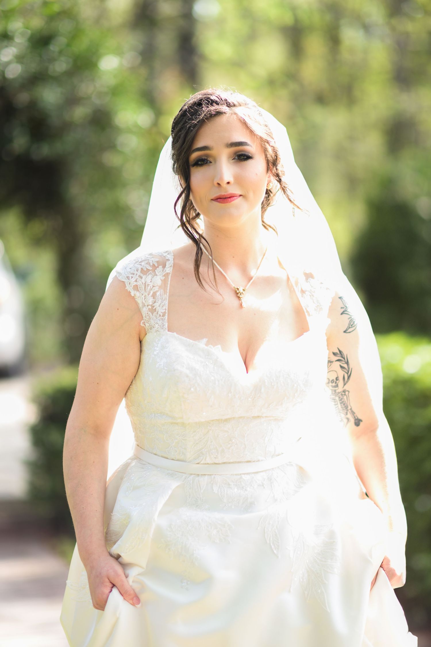 A bride in a white lace wedding dress poses outdoors with soft natural lighting in a garden setting.