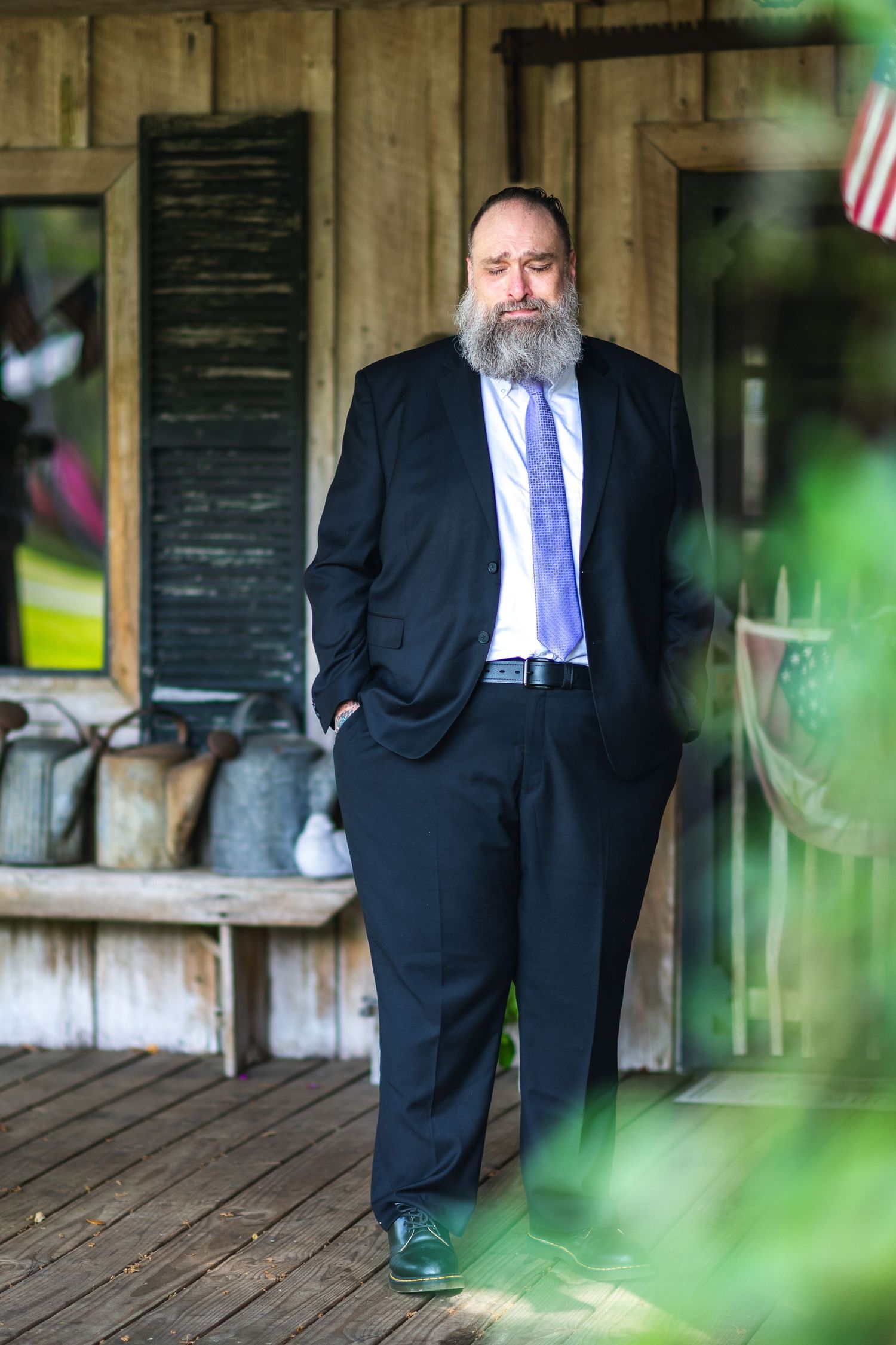 A bearded person in a dark business suit and tie stands on a wooden porch with rustic decor in the background.