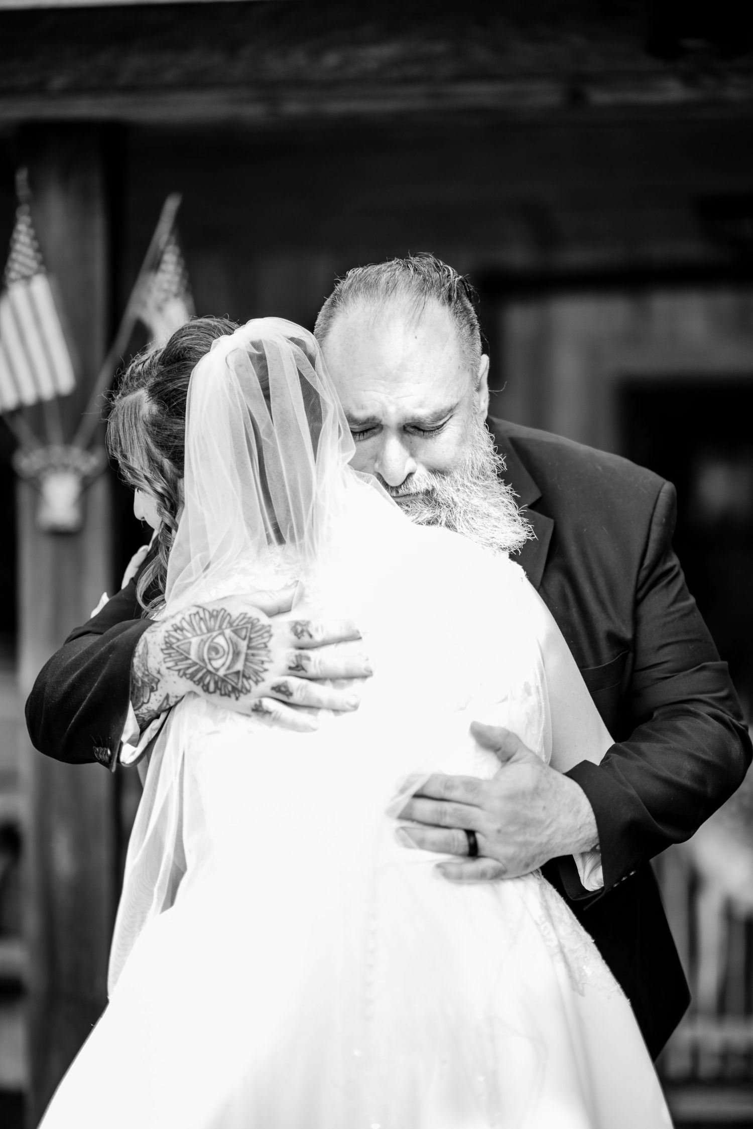 A bride and bearded father share an emotional embrace during a wedding ceremony with an American flag in the background.