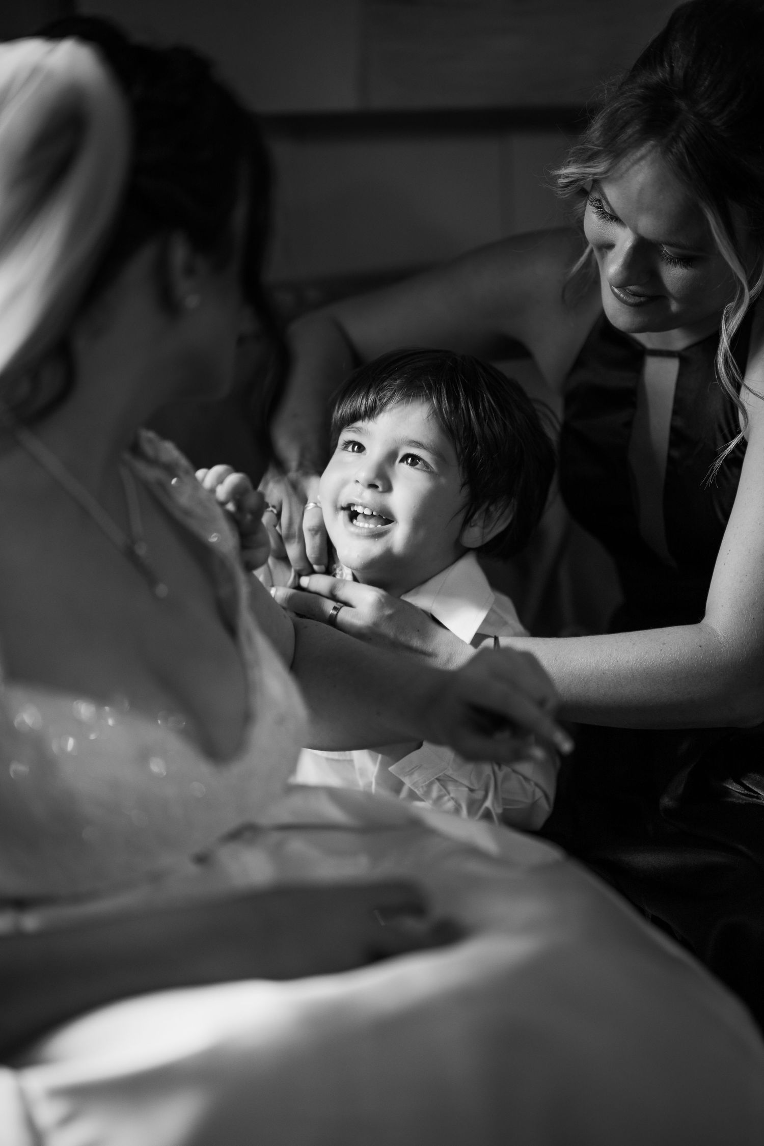Black and white candid photo of a joyful interaction between a child wearing formal attire and adults during a special occasion.