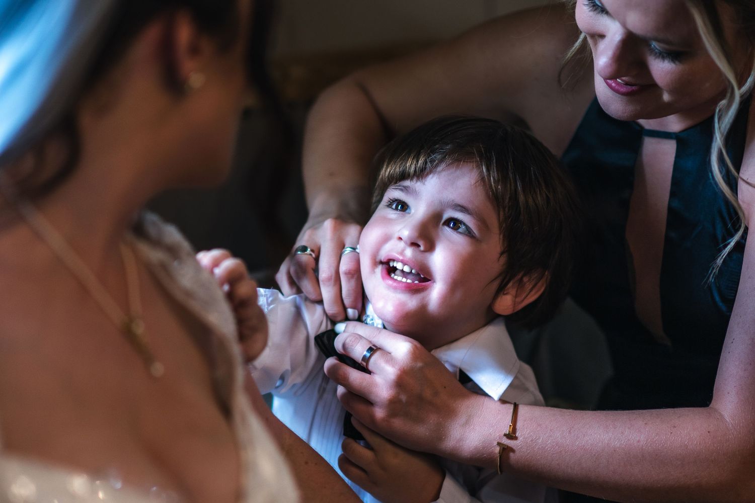 Child wearing a white dress shirt smiles brightly while sitting with others indoors.