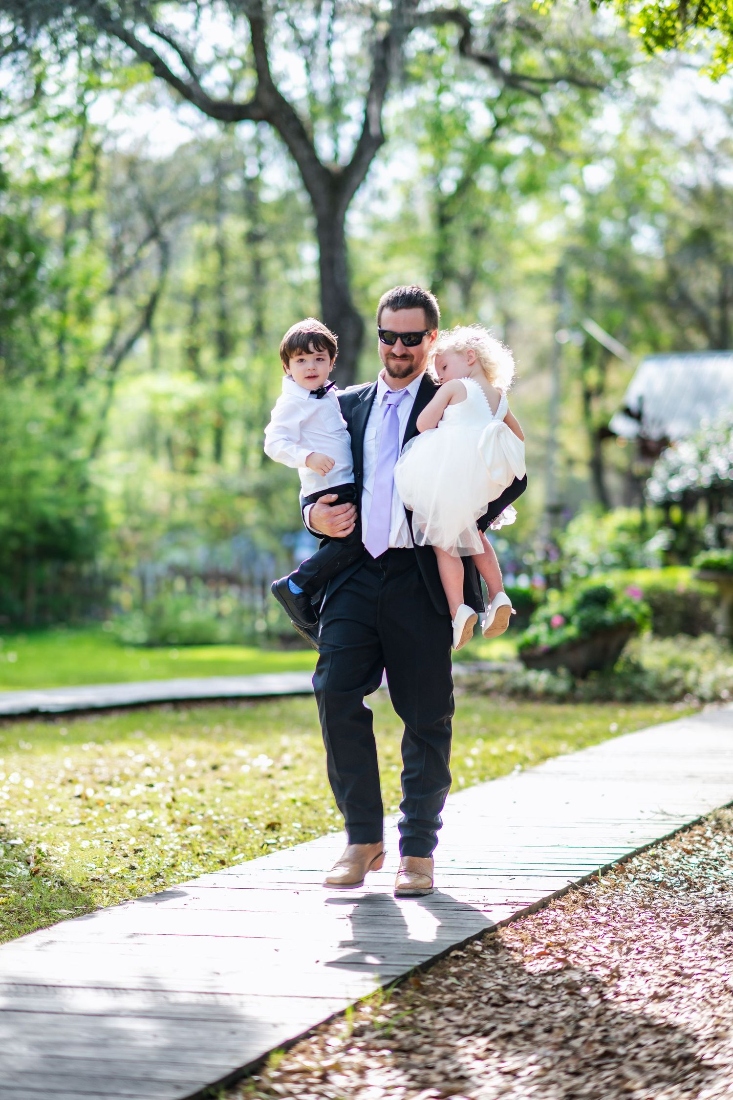 A person stands barefoot on a wooden pathway in a lush garden while holding two small children on a sunny day.