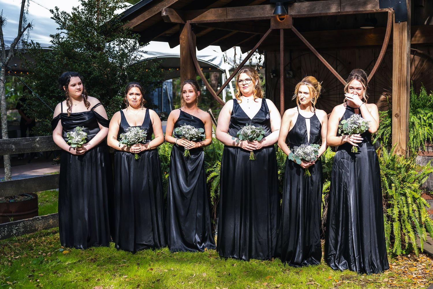 Bridesmaids in elegant black floor-length dresses pose outdoors holding bouquets under a wooden pavilion.