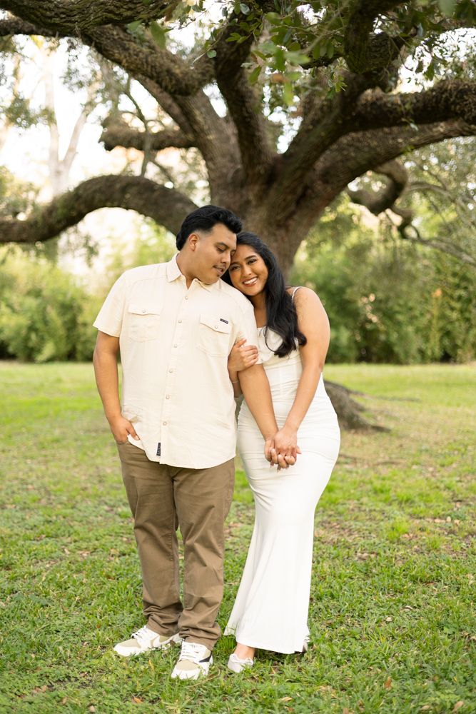 A couple holds hands while standing together in front of a large oak tree in a grassy outdoor setting.
