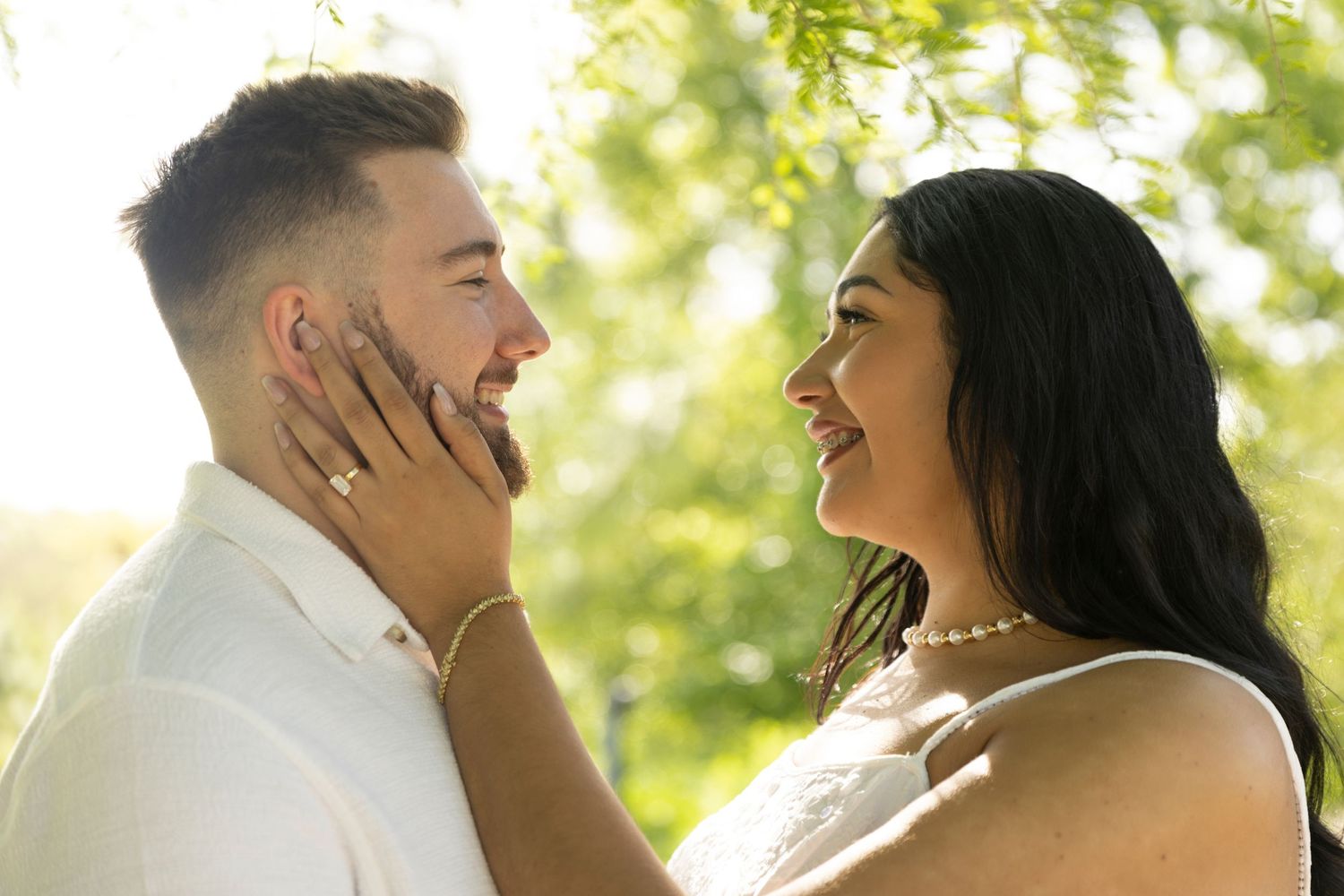 Two people share an intimate outdoor moment in soft sunlit surroundings with trees in background.
