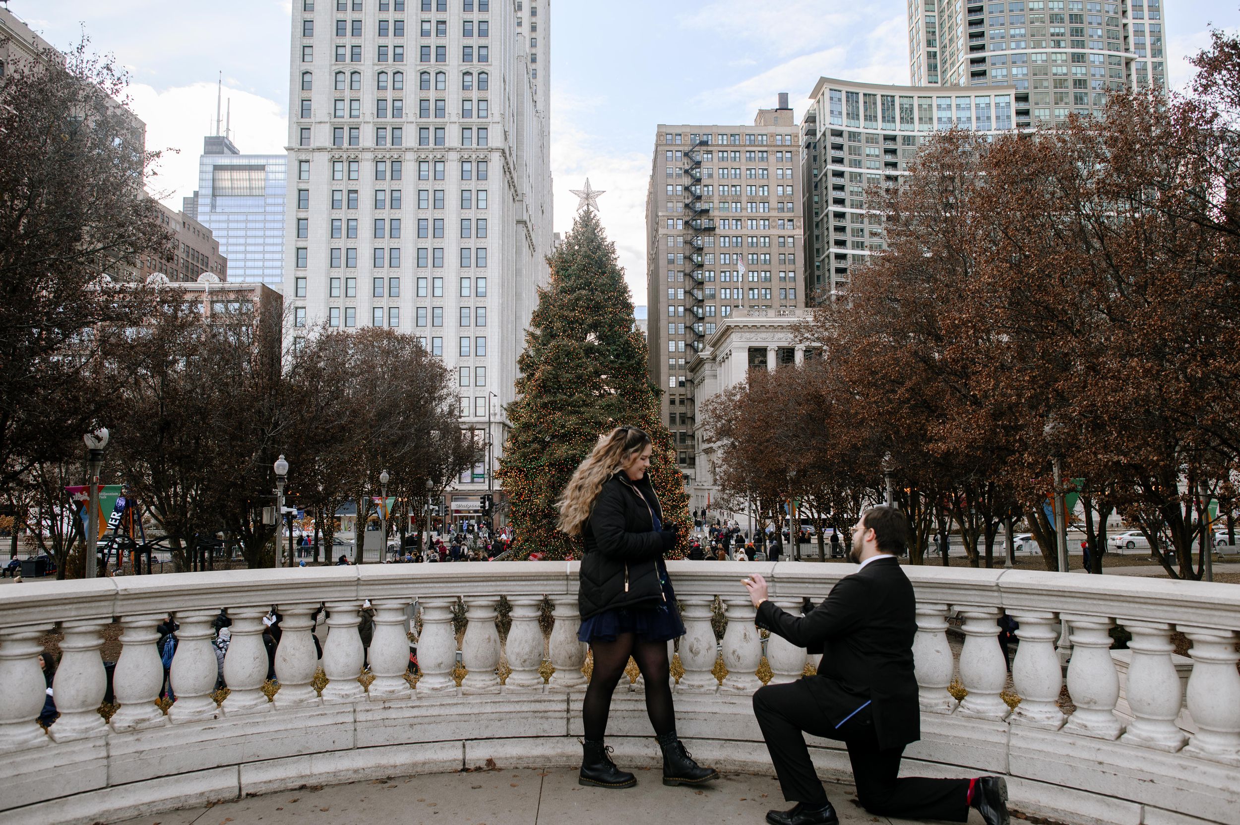 A romantic proposal moment on a balcony overlooking a city park with tall buildings and autumn trees in the background.