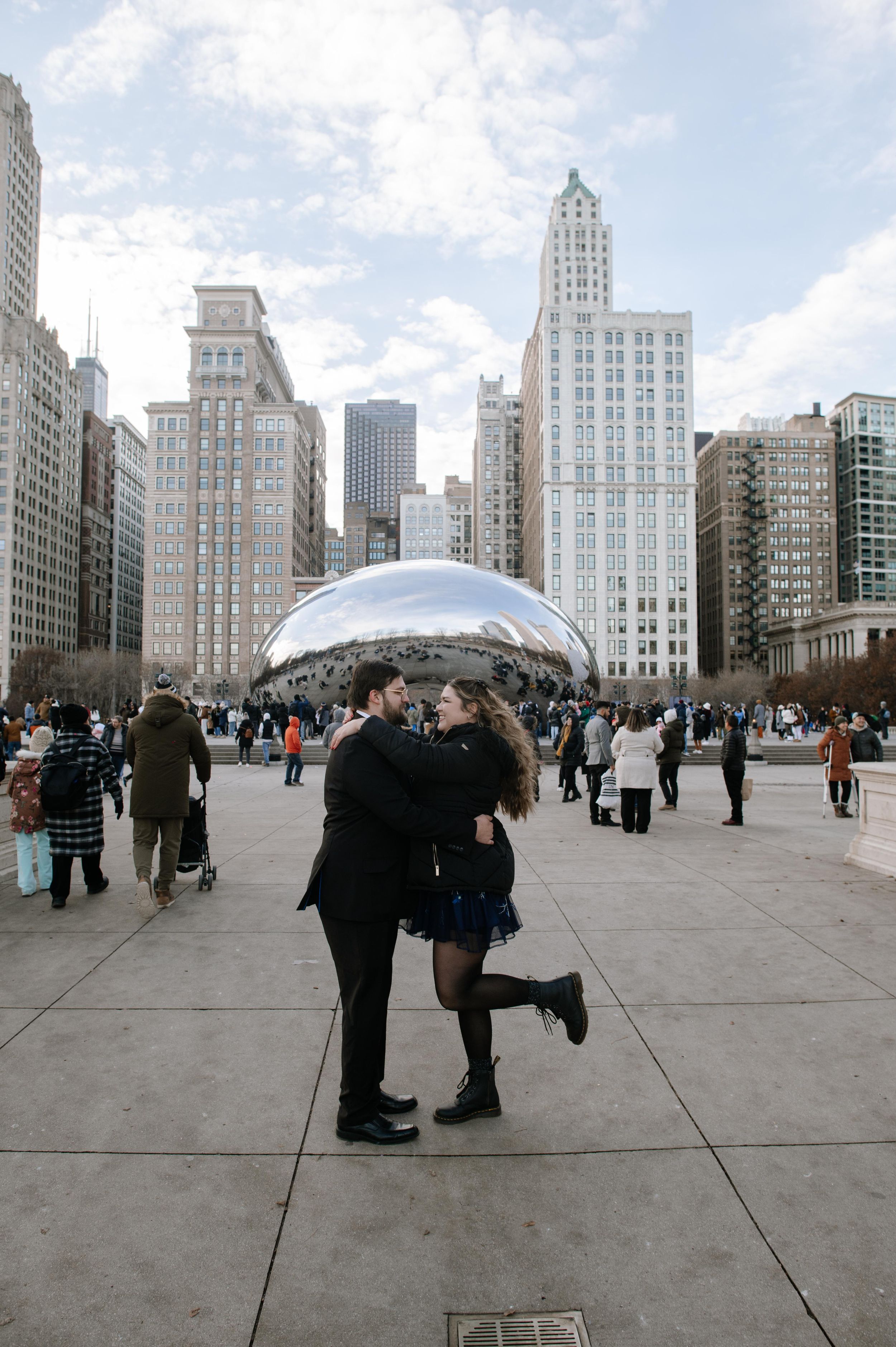 A couple embraces in front of Cloud Gate (The Bean) sculpture with Chicago's iconic skyline and tall buildings in the background.