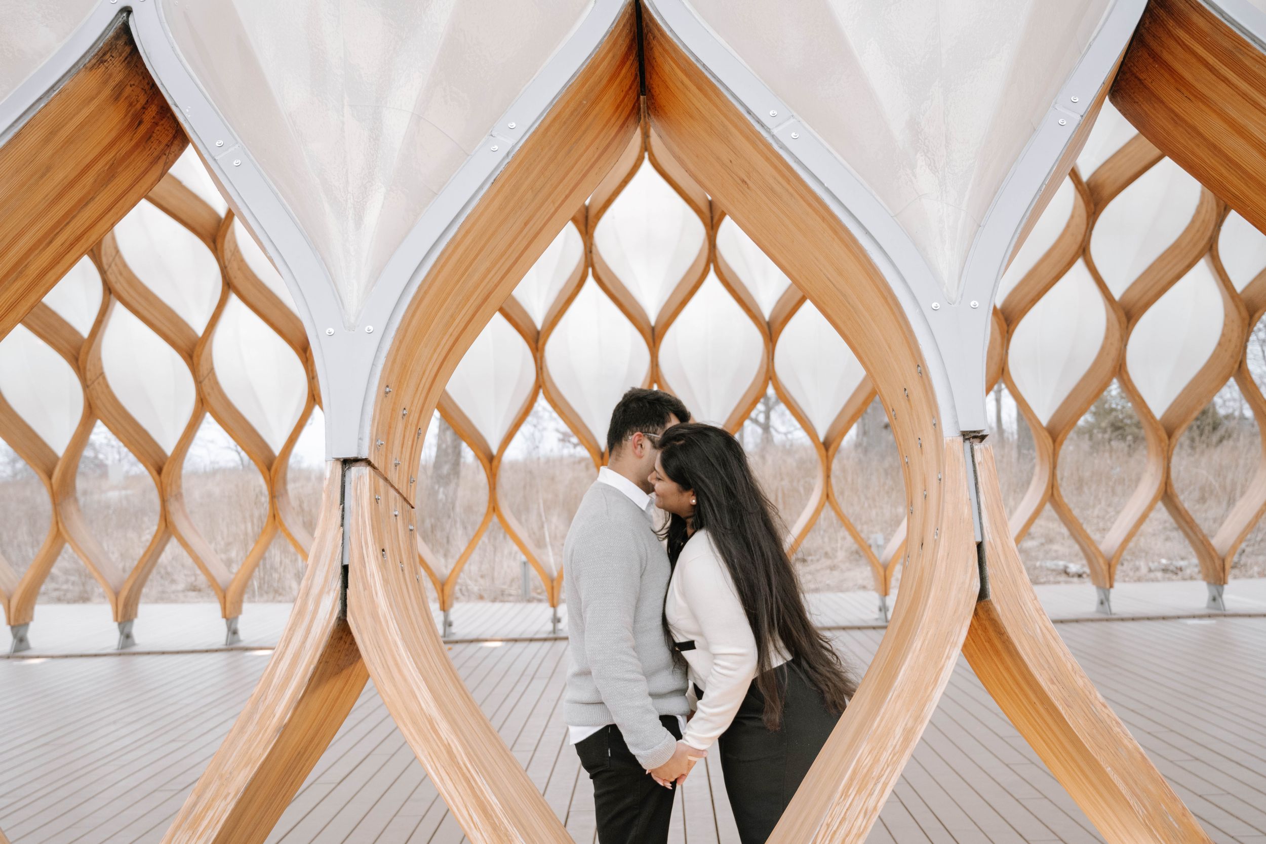 A romantic moment shared between two people framed by a modern wooden architectural structure with diamond lattice patterns.