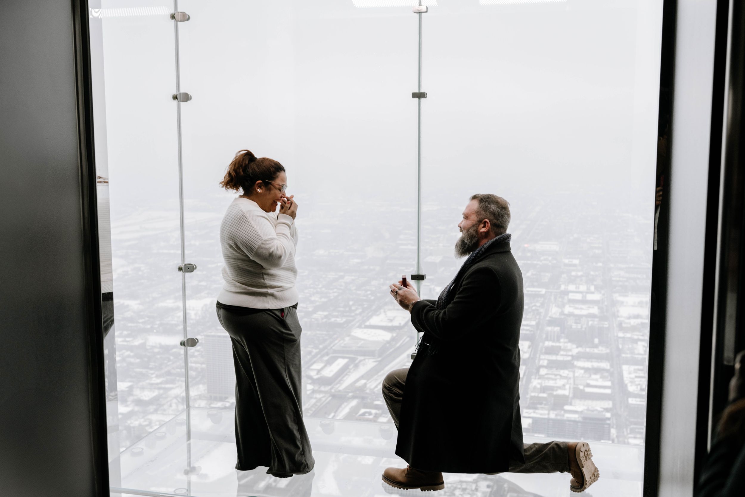 A romantic proposal takes place at a skyscraper observation deck with a misty cityscape view in the background.