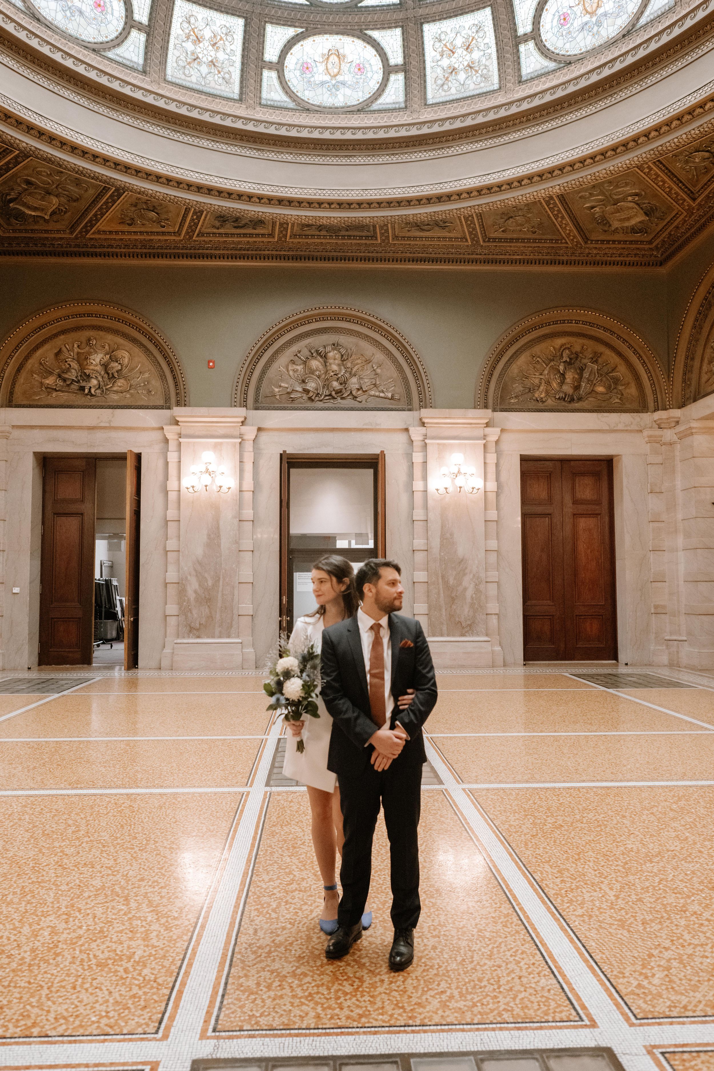 A couple stands in an ornate circular room with marble floors, decorative ceiling, and elegant architectural details.