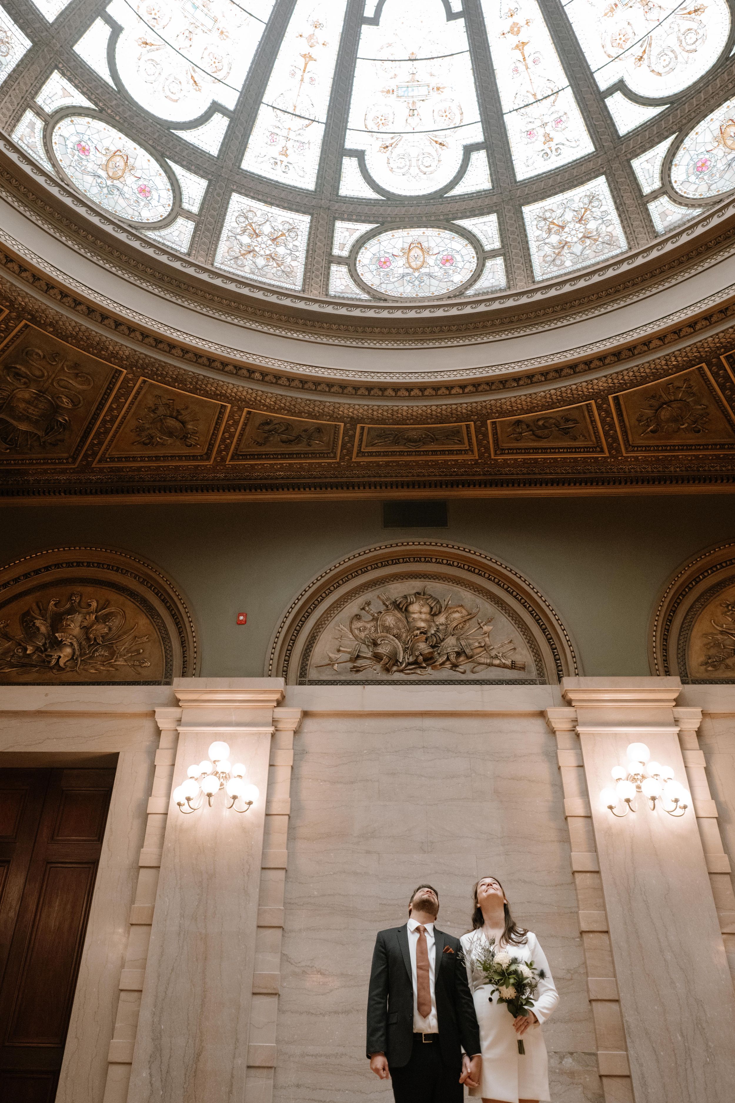 Elegant couple stands beneath ornate domed ceiling with decorative details and crystal sconces in grand historic building.