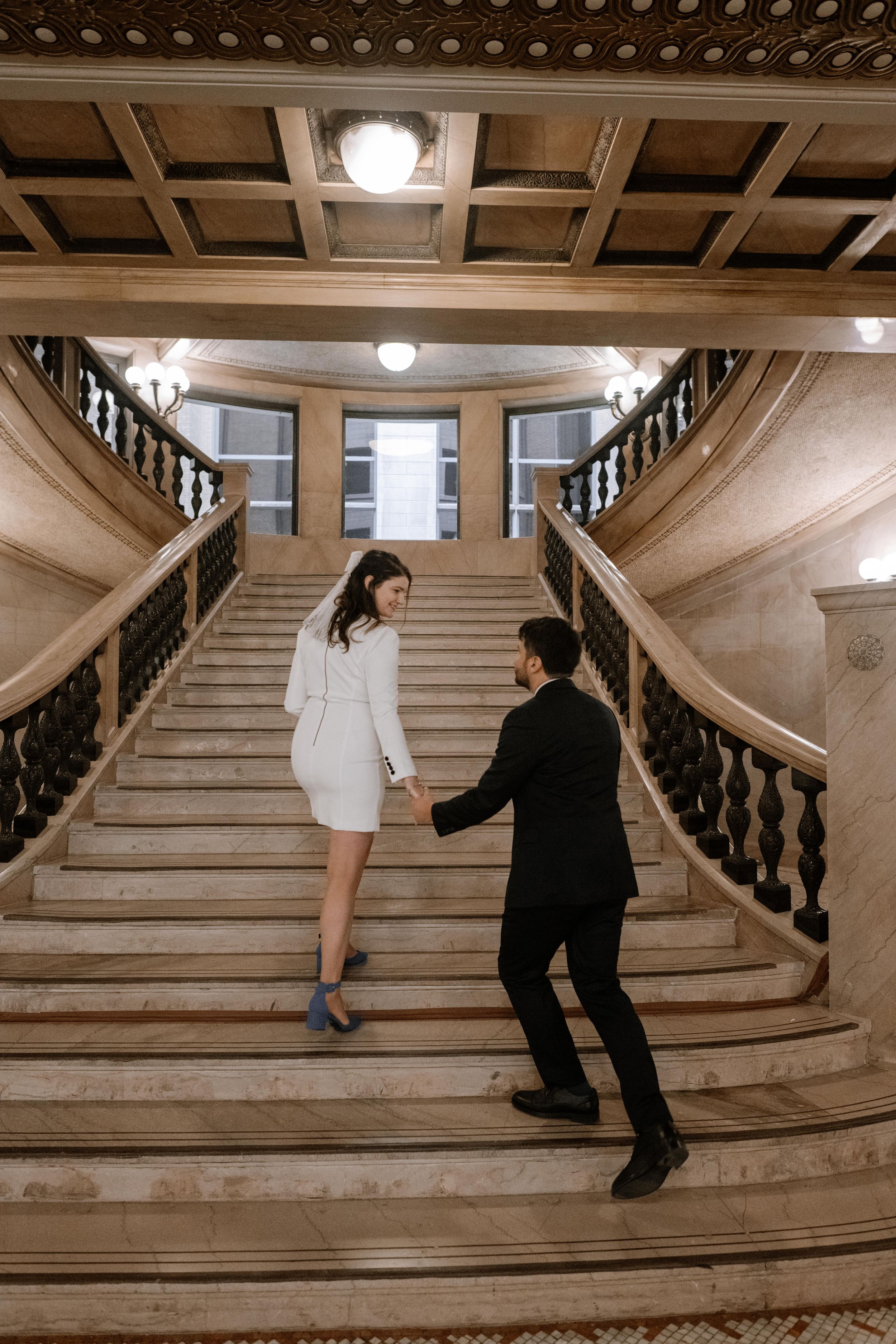 Two people in contrasting black and white outfits pose on an elegant marble staircase with ornate railings and coffered ceiling.