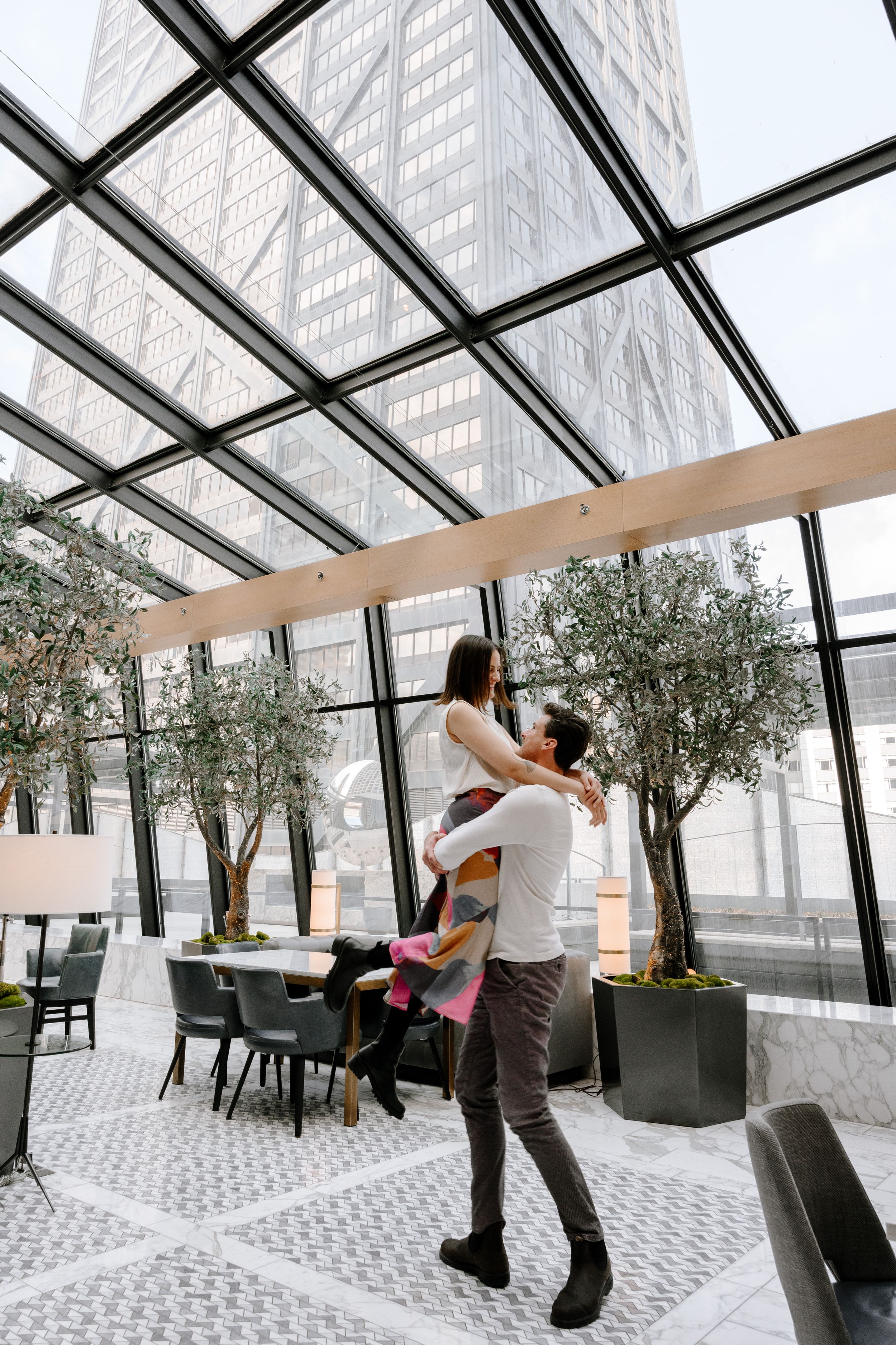 Romantic couple embraces in a modern glass-roofed restaurant decorated with olive trees and geometric tile flooring.