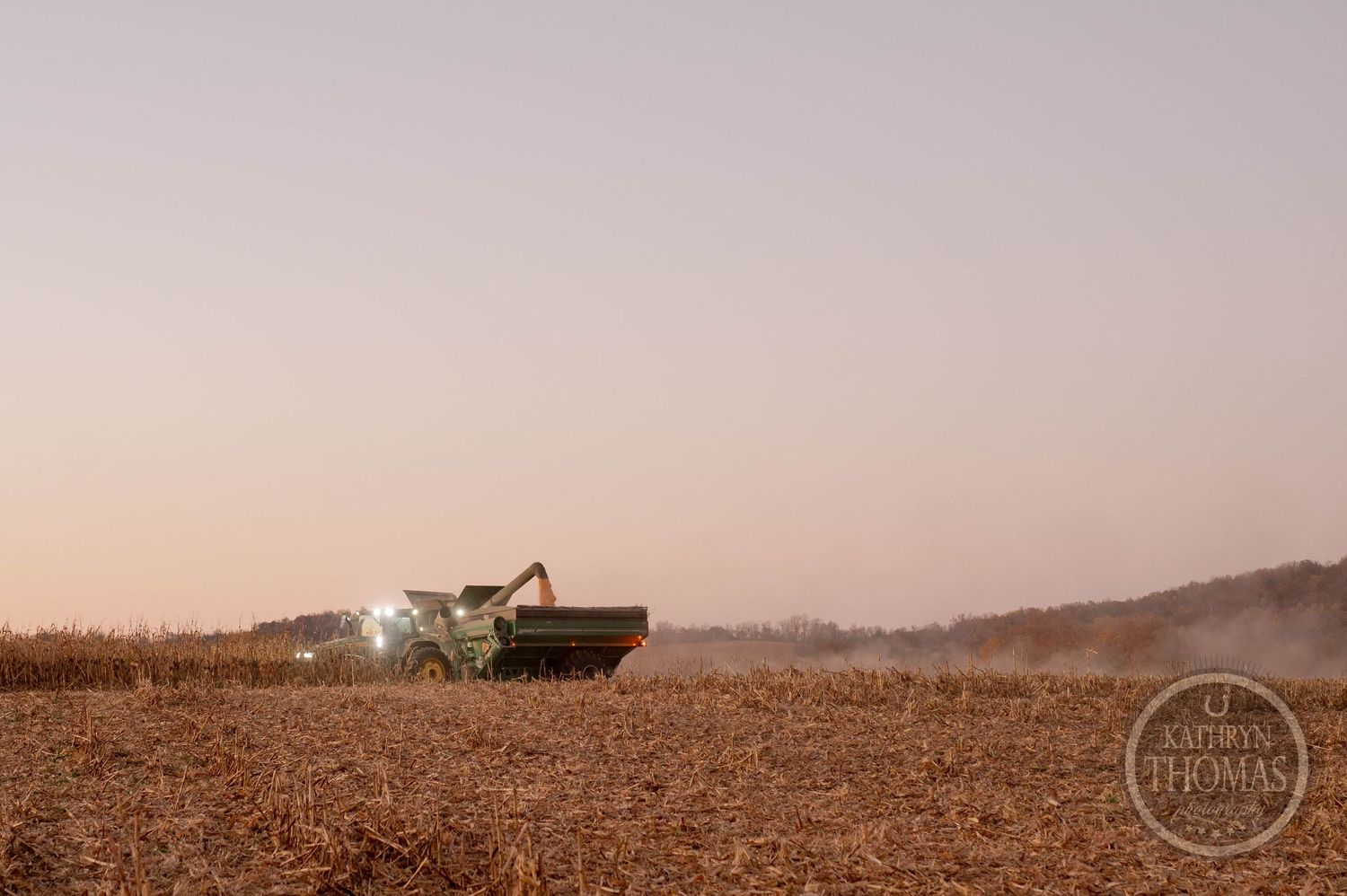 Farm equipment operating in a dusty field at sunset with lights glowing.