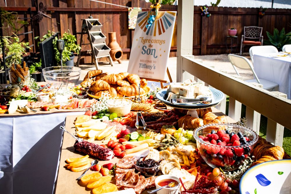 Elegant charcuterie board display with meats, cheeses, fruits and snacks arranged on white tablecloths at an outdoor event.