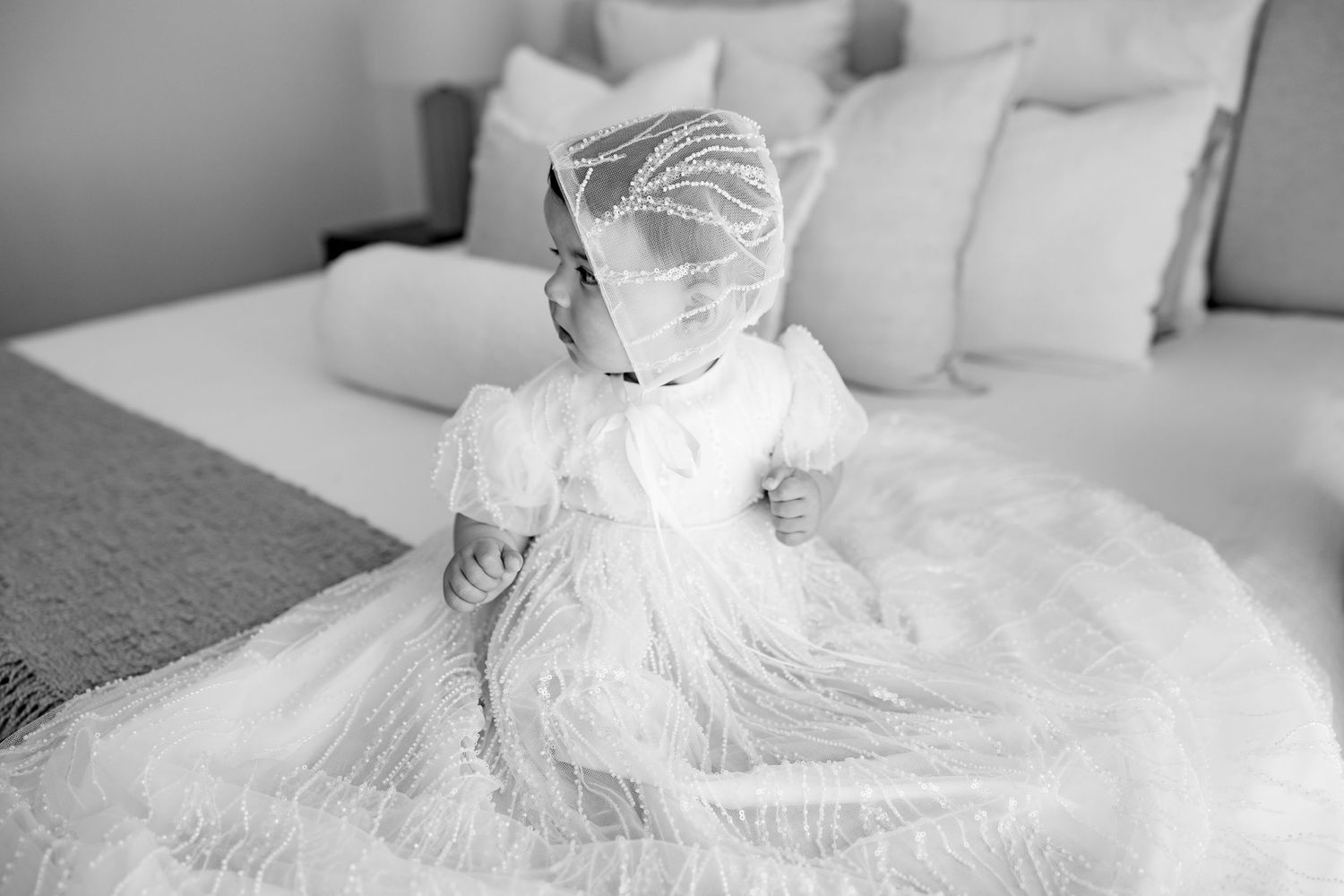 White baptismal dress spread out on bed with soft natural lighting in monochrome style.