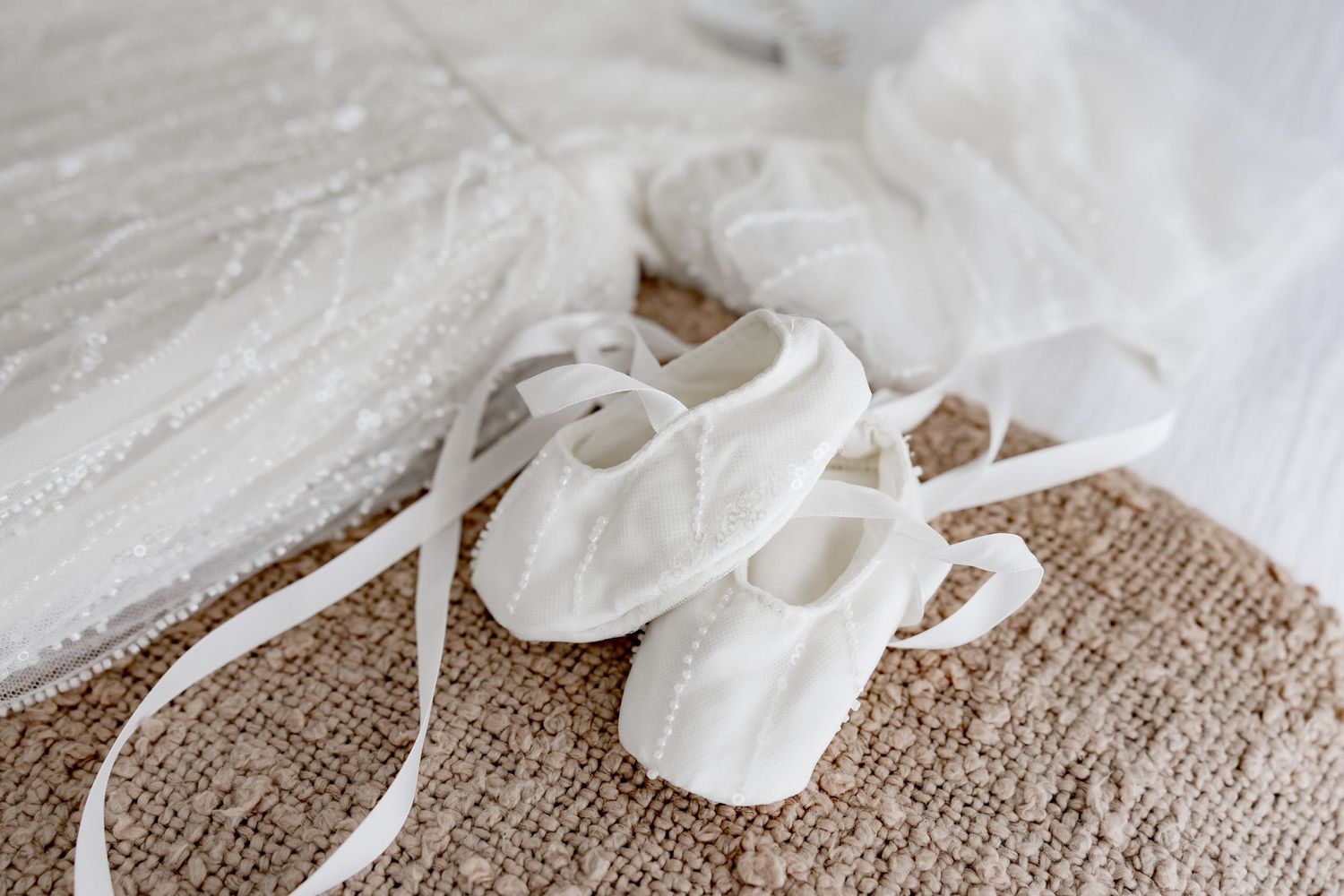 Tiny white baby shoes with satin ribbons displayed on a textured beige fabric surface.