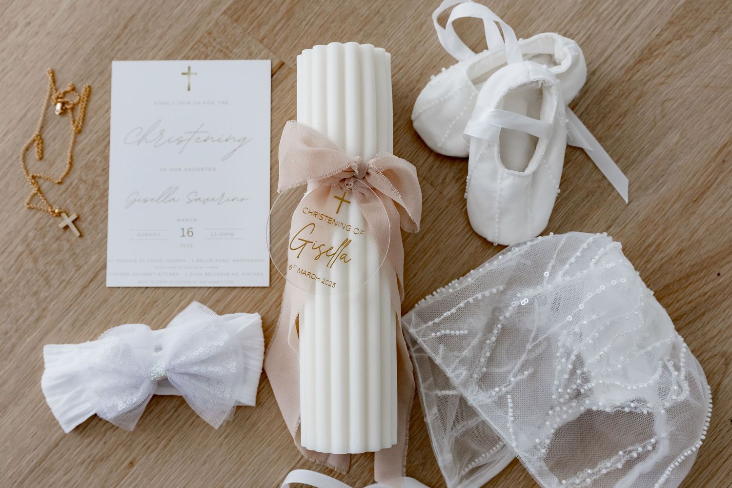 A collection of white christening items including a candle, booties, bow and lace veil laid on a wooden surface.