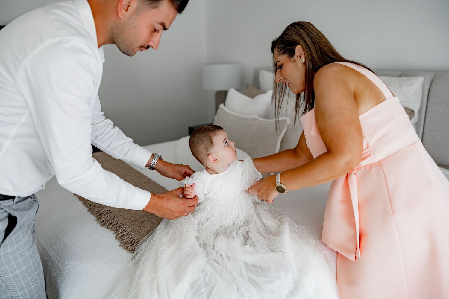 Two people help dress a baby in a white christening gown on a bed.