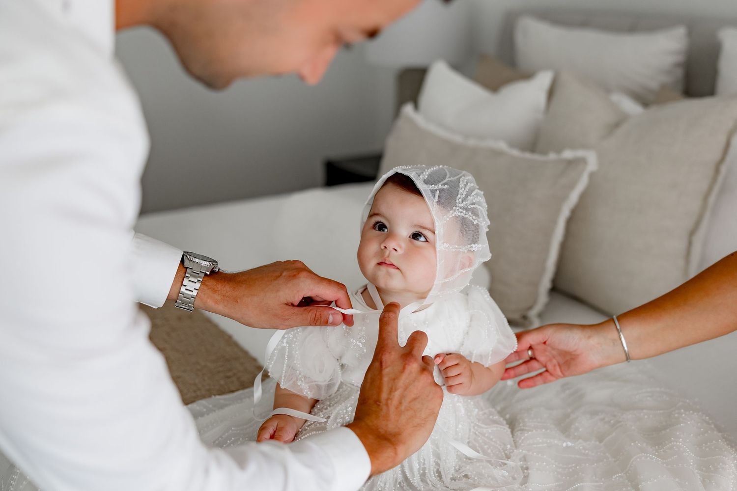 A baby in a white christening gown and bonnet being carefully dressed on a light gray bed.