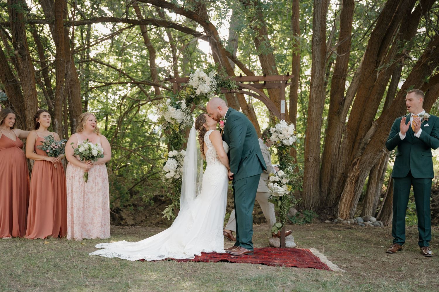 Wedding party stands in a line under a canopy of trees in an outdoor garden setting.