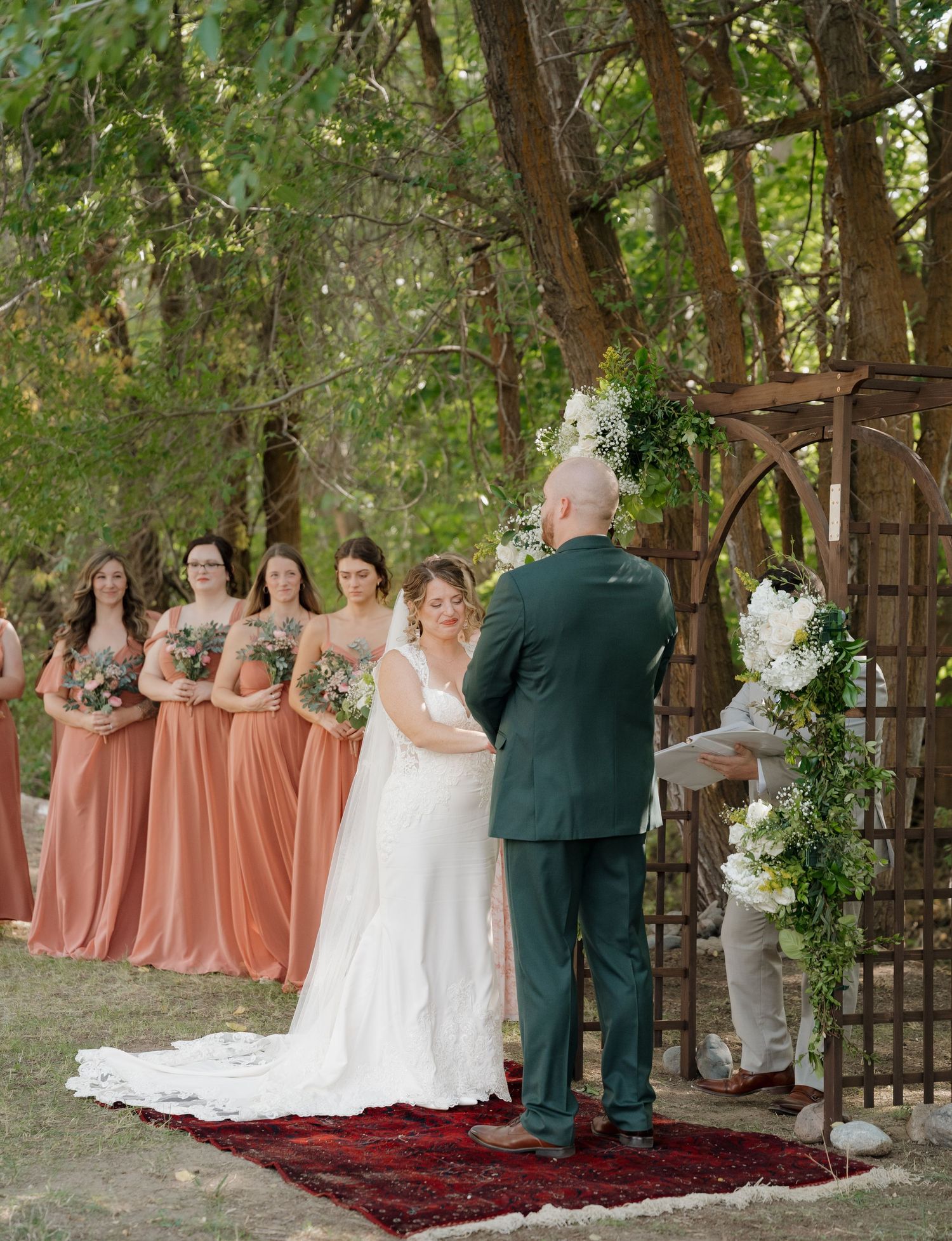 Wedding ceremony setup in a wooded outdoor setting with bridesmaids in coral dresses and a decorative arch.