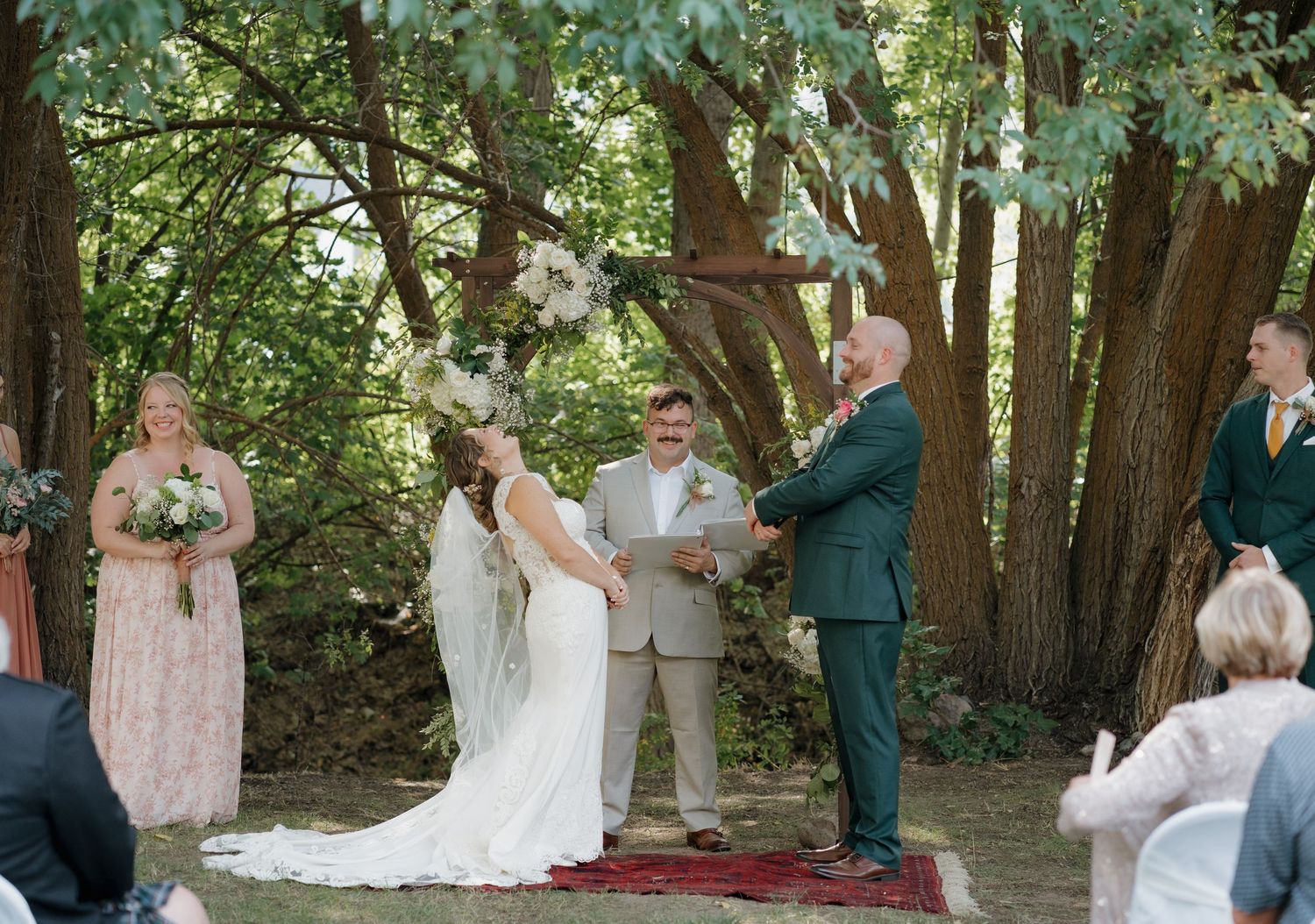 Panoramic view of an outdoor wedding ceremony in a wooded setting with guests gathered under tall trees.