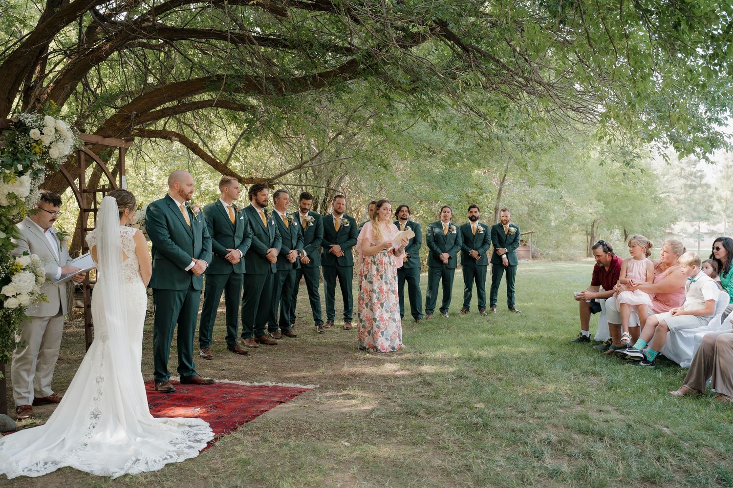 Wedding ceremony participants stand in line under green trees during an outdoor summer celebration.