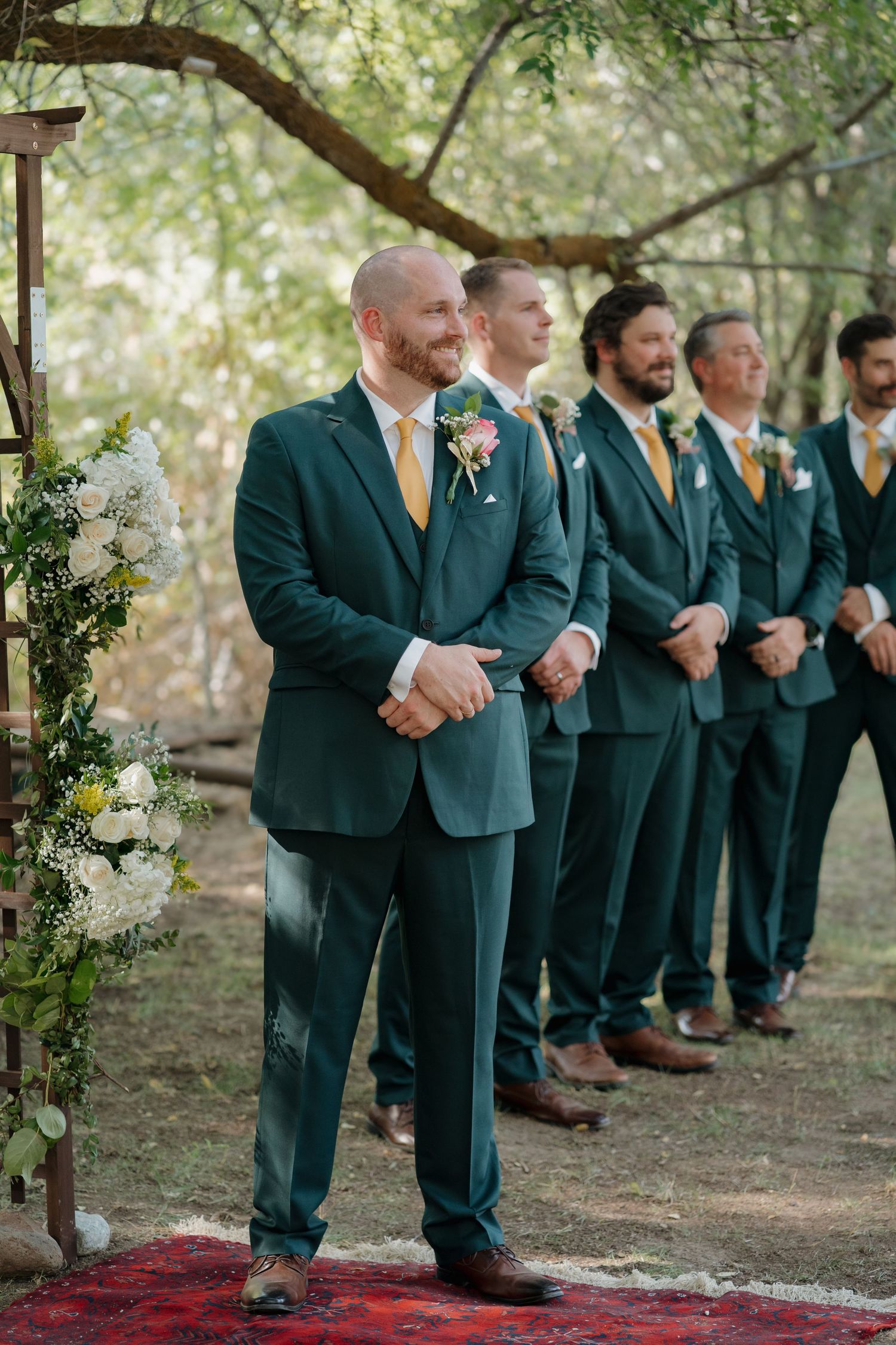Groomsmen in emerald green suits with yellow ties stand in line at an outdoor wedding ceremony under trees.