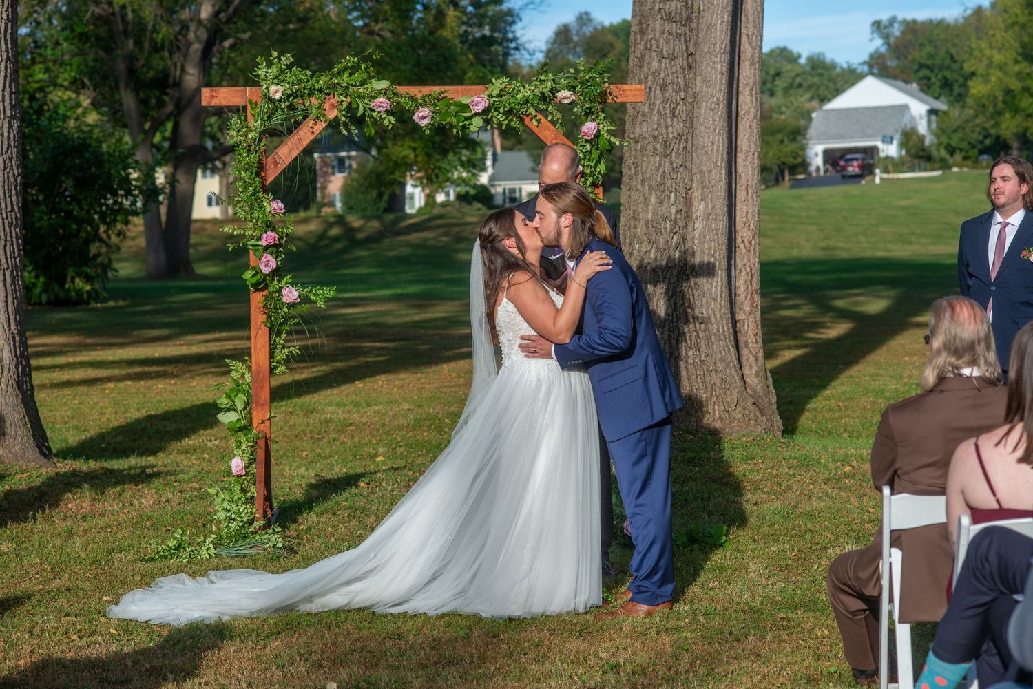 Wedding party poses for photos in a sunny outdoor garden setting with trees and wooden decor elements.