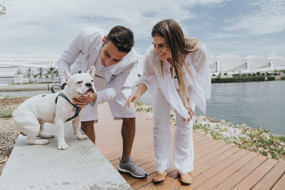 A couple in white outfits plays with their white dog on a waterfront wooden deck during a sunny day.