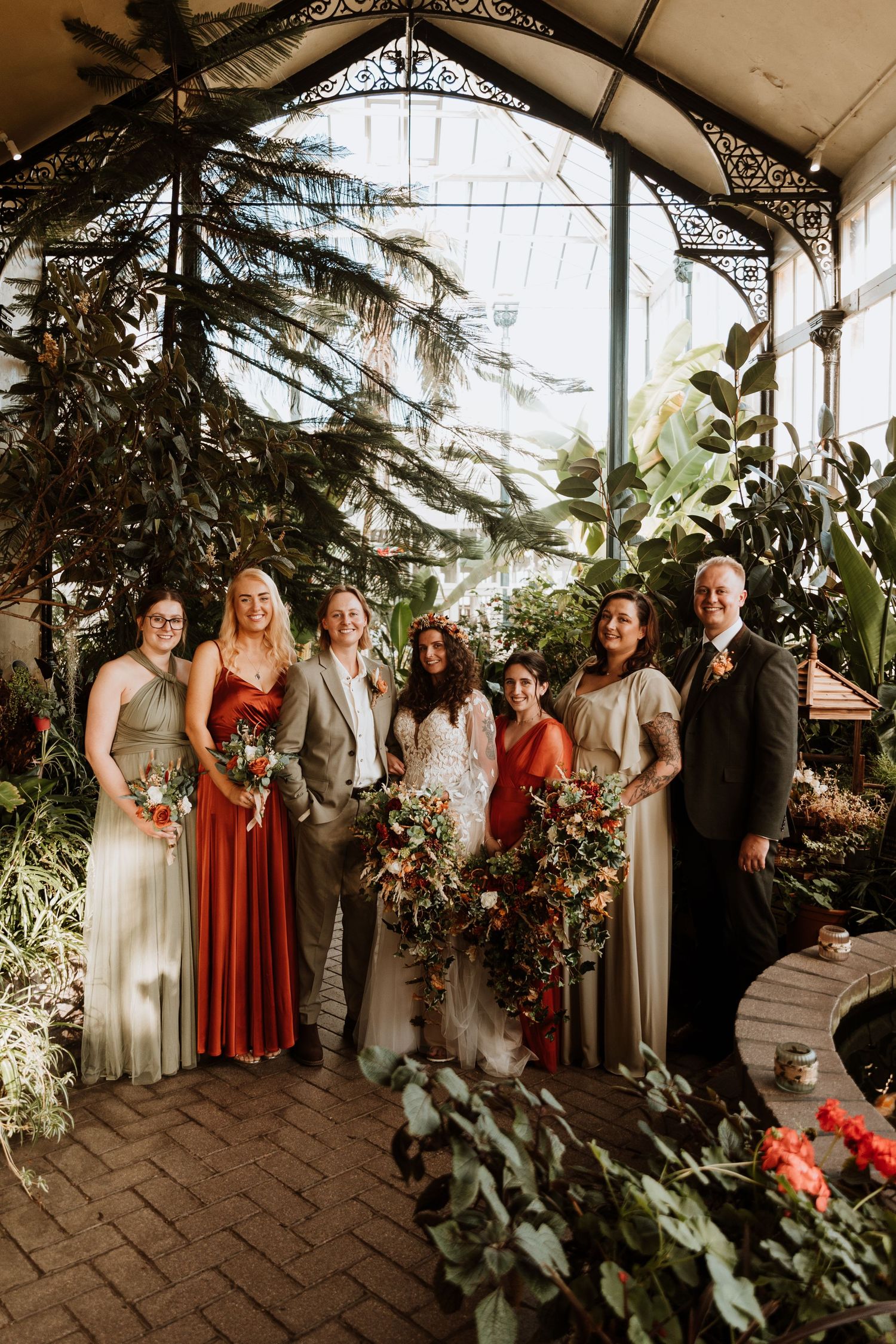 Wedding party poses in a greenhouse conservatory with lush tropical plants and natural light filtering through glass panels.