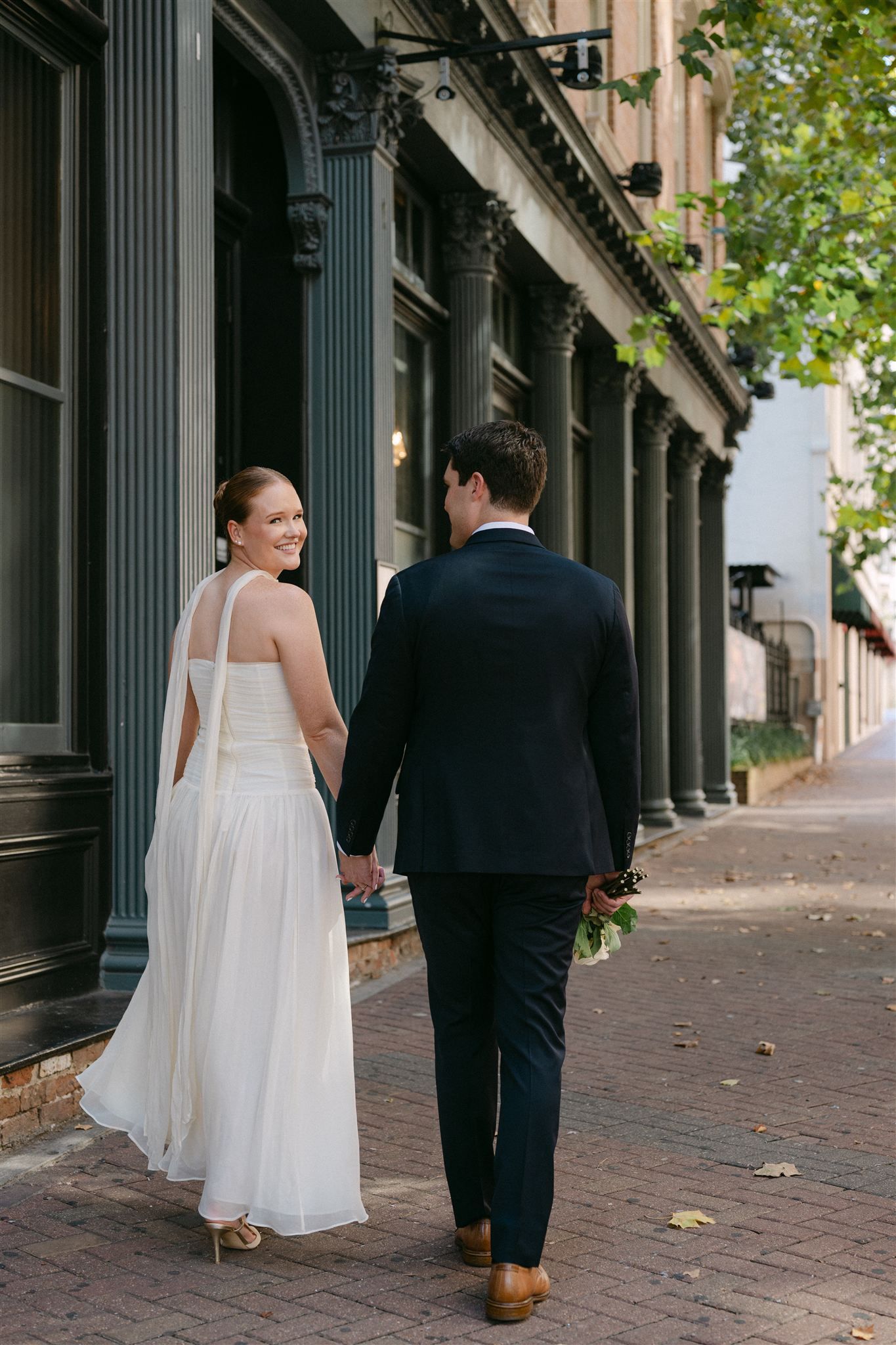 A bride and groom walk along a historic brick sidewalk with elegant iron columns during their wedding day.