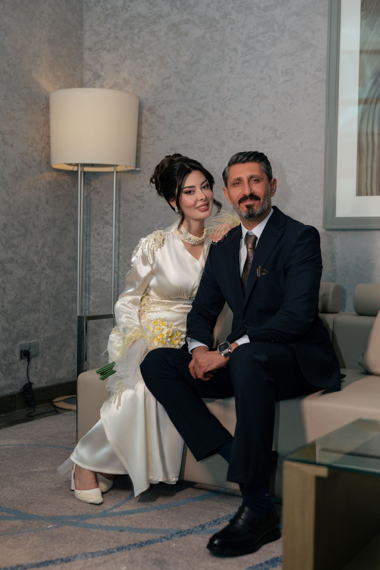 A couple in formal attire sits together on a couch in an elegant room with gray walls and modern decor.