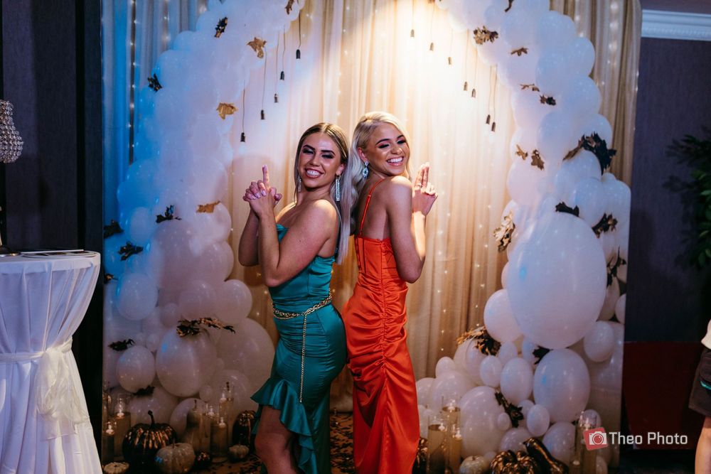 Two people in colorful dresses dance and pose together in front of a butterfly-decorated backdrop at an event.