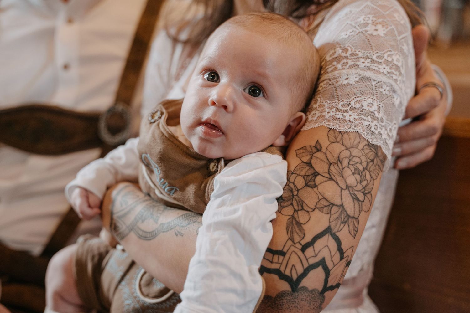 A baby in a white onesie rests against a tattooed arm while looking at the camera with bright eyes.