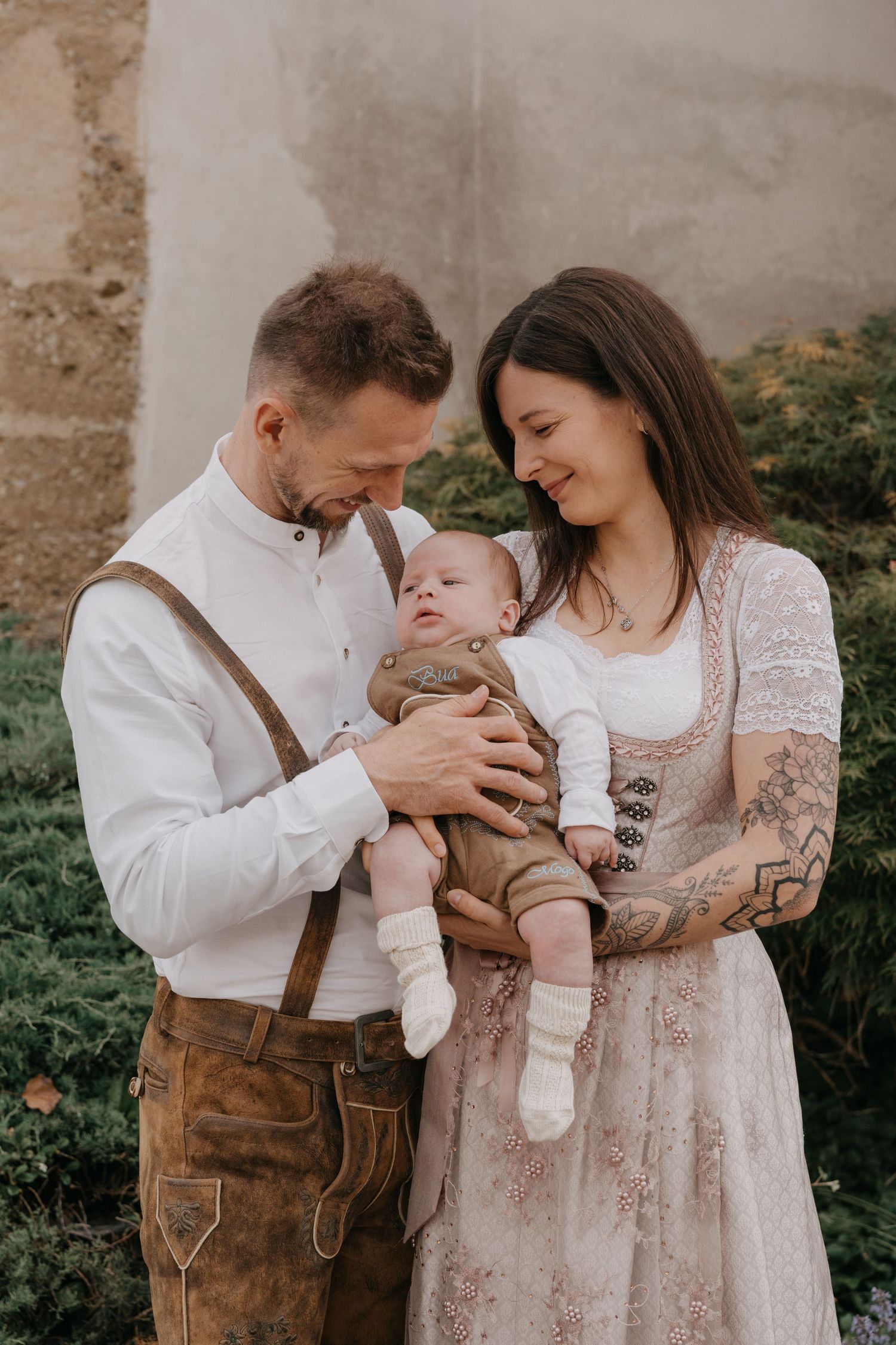 A couple in rustic attire shares tender moments with their baby against a stone wall backdrop in this photo series.