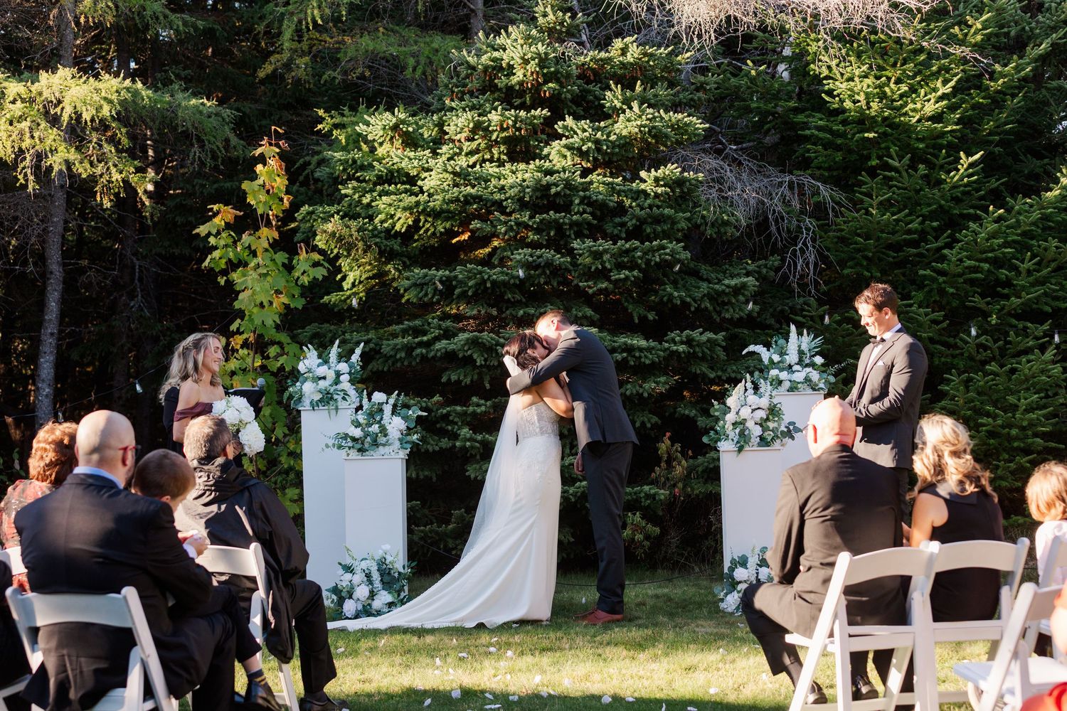 An outdoor garden wedding ceremony takes place at dusk with white floral arrangements and folding chairs for guests.