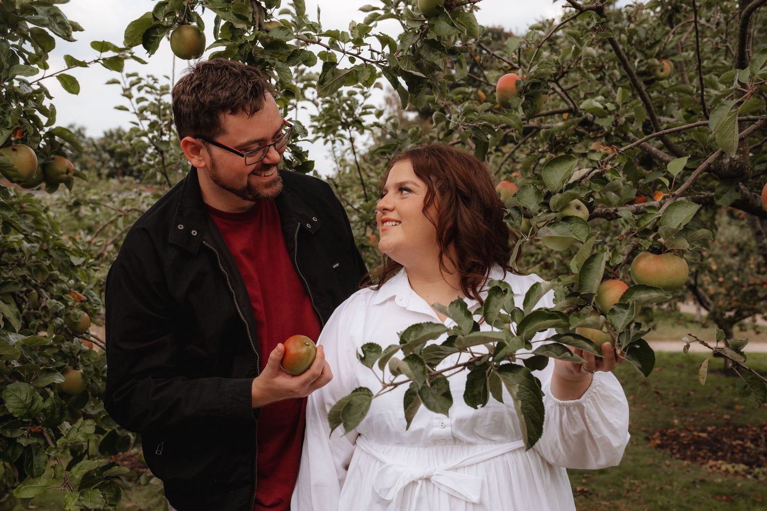Couple smiling at each other while picking apples in a lush green orchard on a cloudy day.
