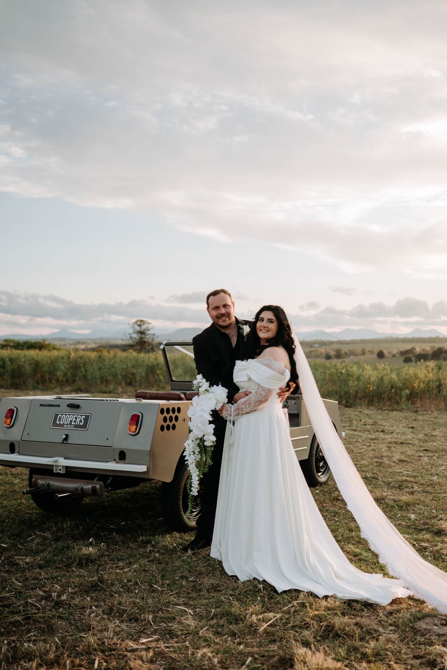A wedding party stands outdoors with vintage cars against a scenic mountain backdrop on a sunny day.