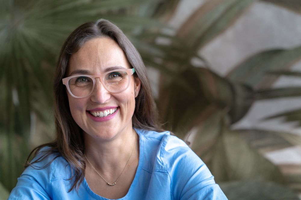 Person wearing glasses and blue top smiles warmly in office environment.