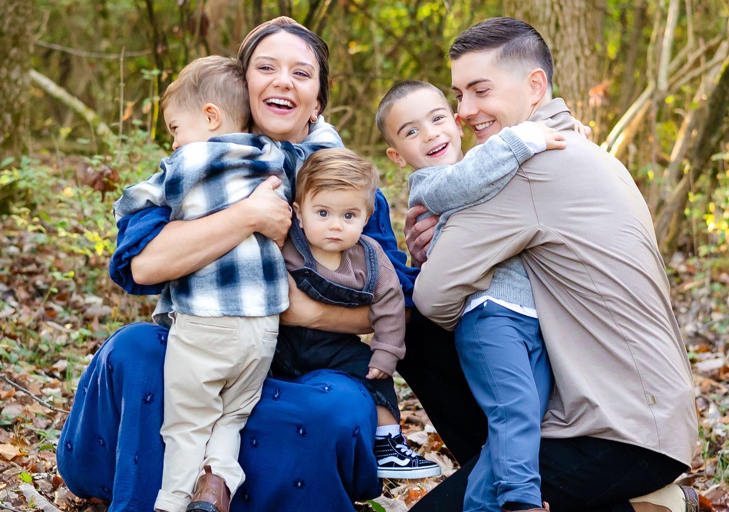A family shares joyful embraces while kneeling together on autumn leaves in a sunlit wooded area.